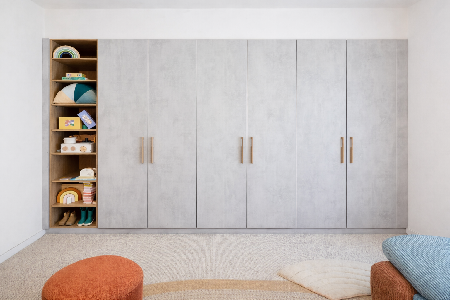 A large modern wardrobe with several grey doors and a wooden open shelving unit on the left, containing children's toys, books, and decorations. The room has a beige carpet, a round orange cushion, a cream-colored cushion, and a light blue cushion on