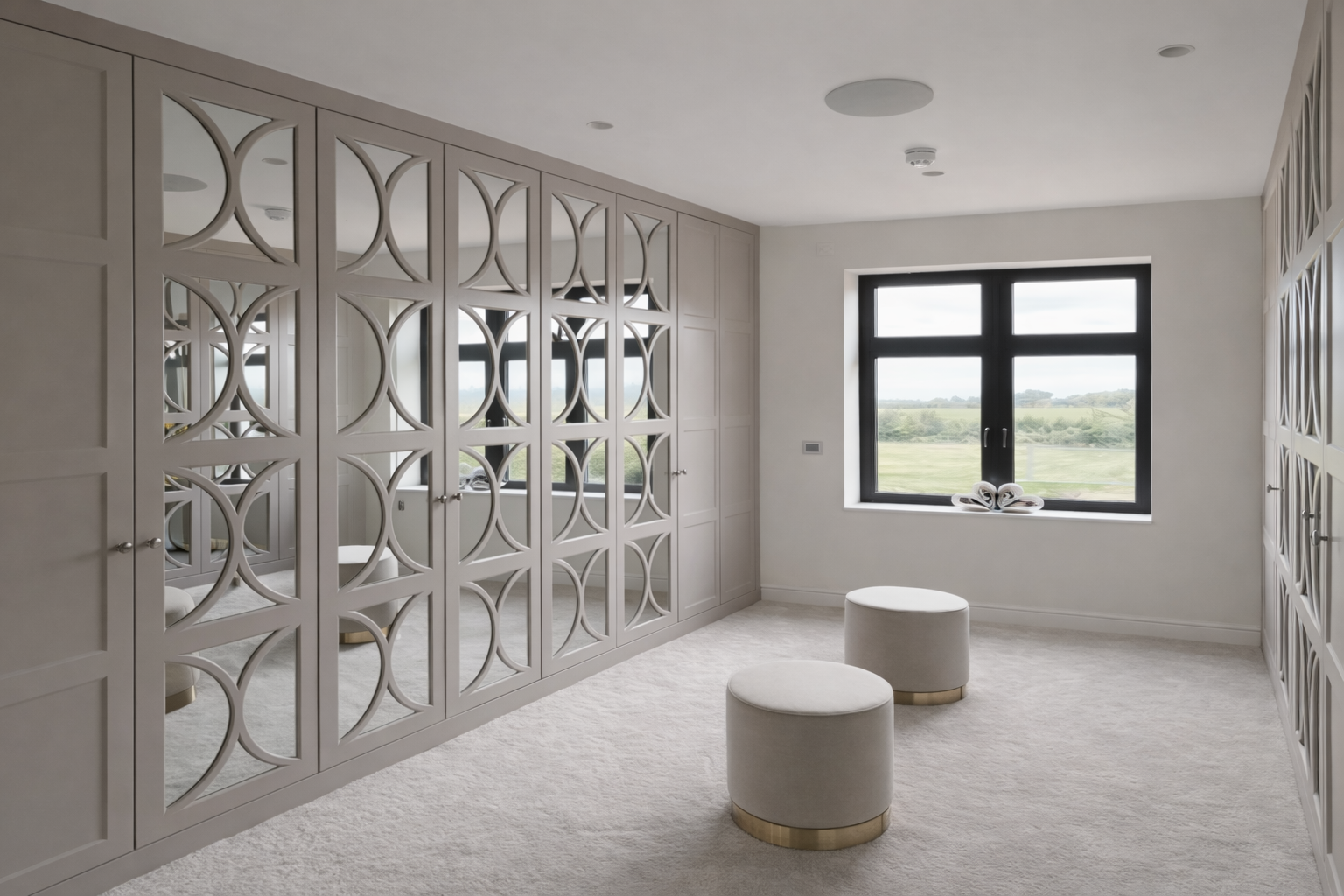 A modern, minimalist room with white carpet, patterned built-in wardrobes with mirrors, a large window showing a green landscape, and two white ottomans with gold bases.