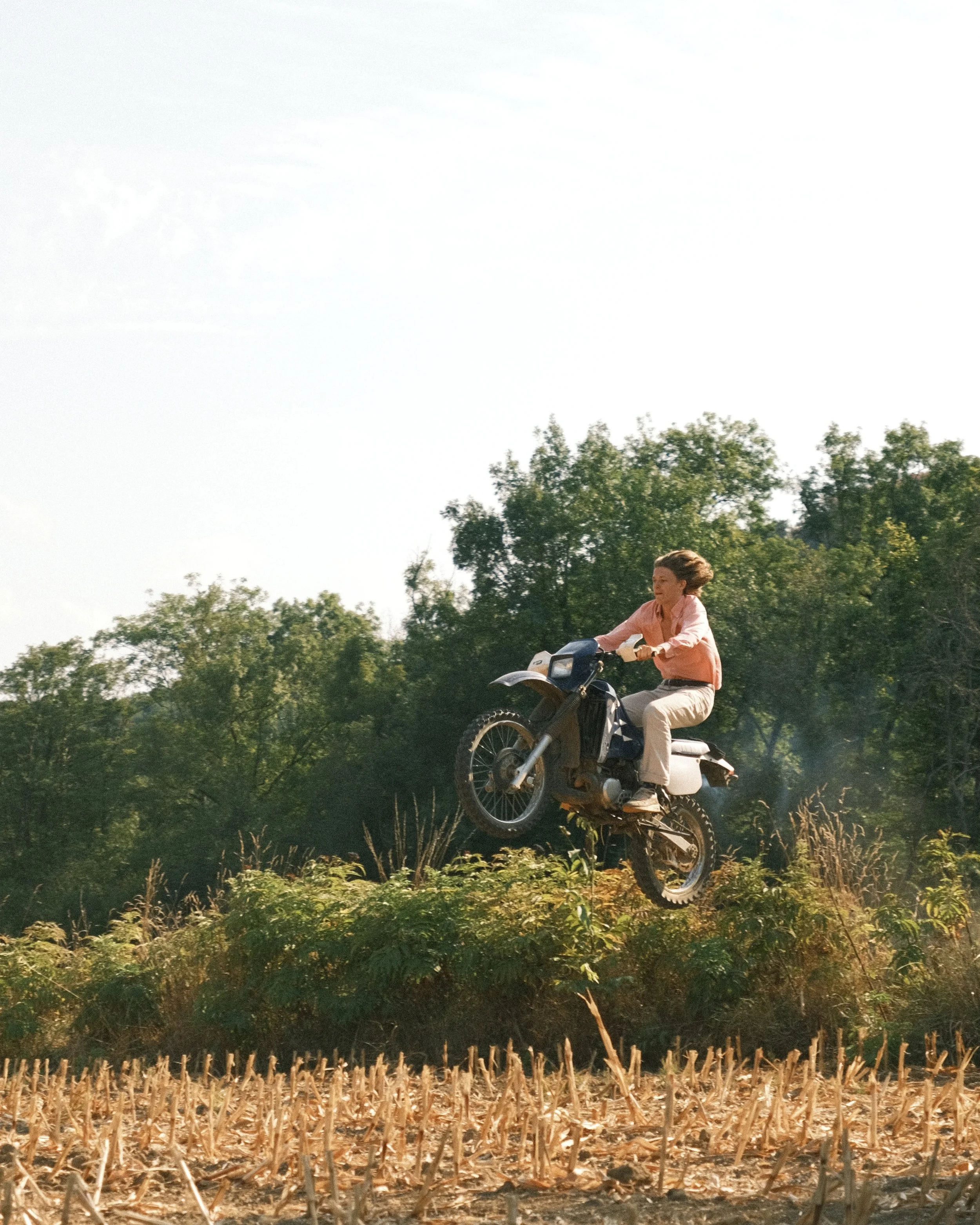 Une femme en rose clair, portant un pantalon beige, sautes en moto dans un champ avec des arbres en arrière-plan et un ciel clair.