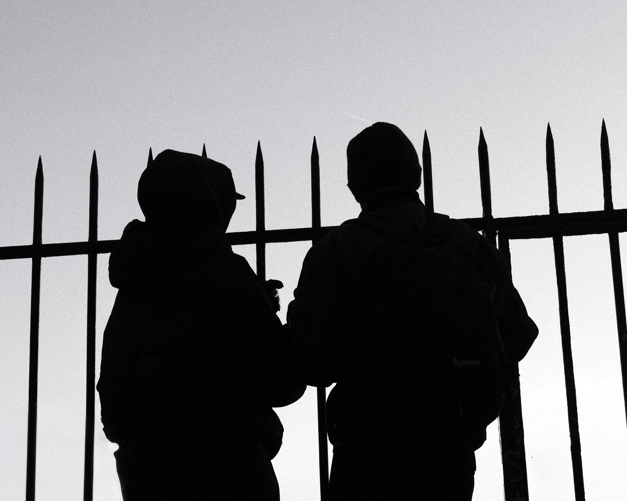 Silhouettes de deux personnes devant une clôture en fer forgé, en arrière-plan un ciel clair.