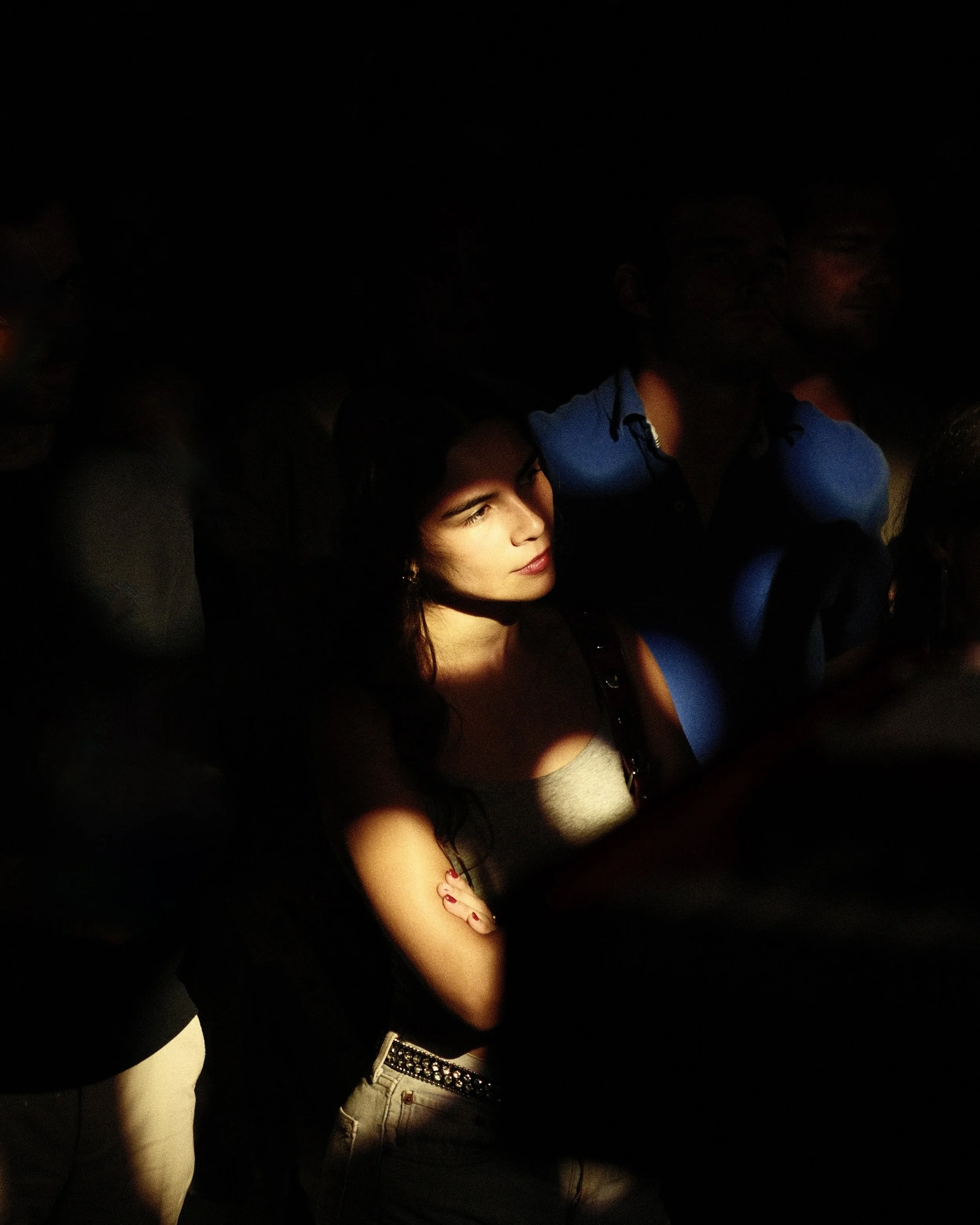 Une femme regarde vers la gauche, éclairée par un faisceau de lumière dans un fond sombre, entourée de plusieurs personnes dans l'ombre.