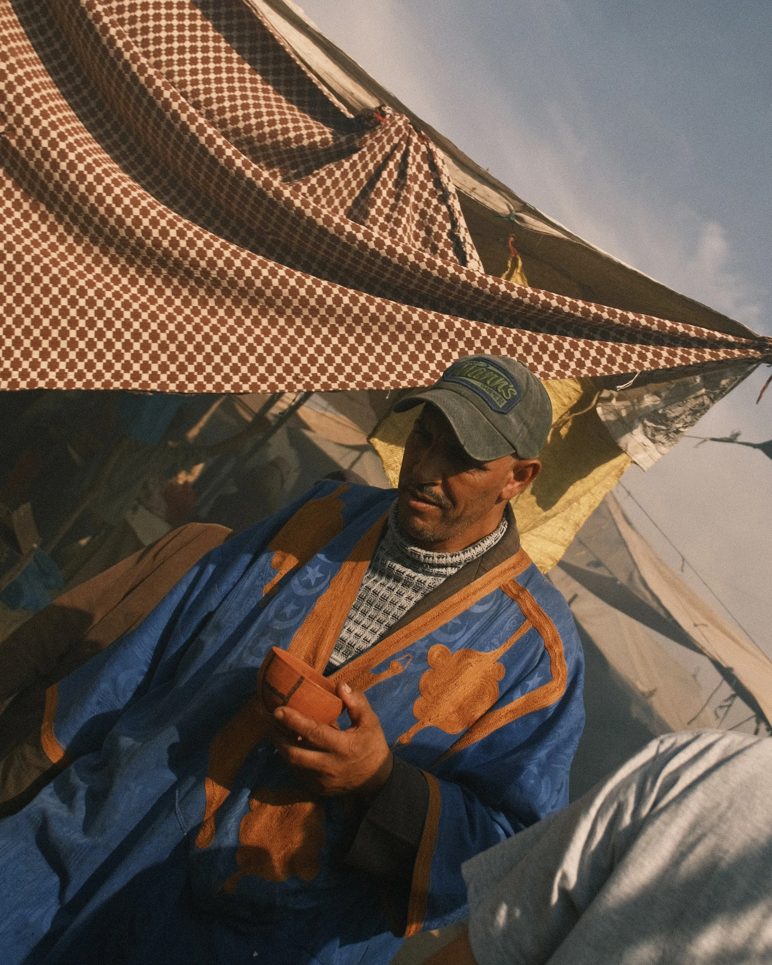 Un homme portant une casquette et une veste colorée, sous une tente avec un fond de ciel clair.