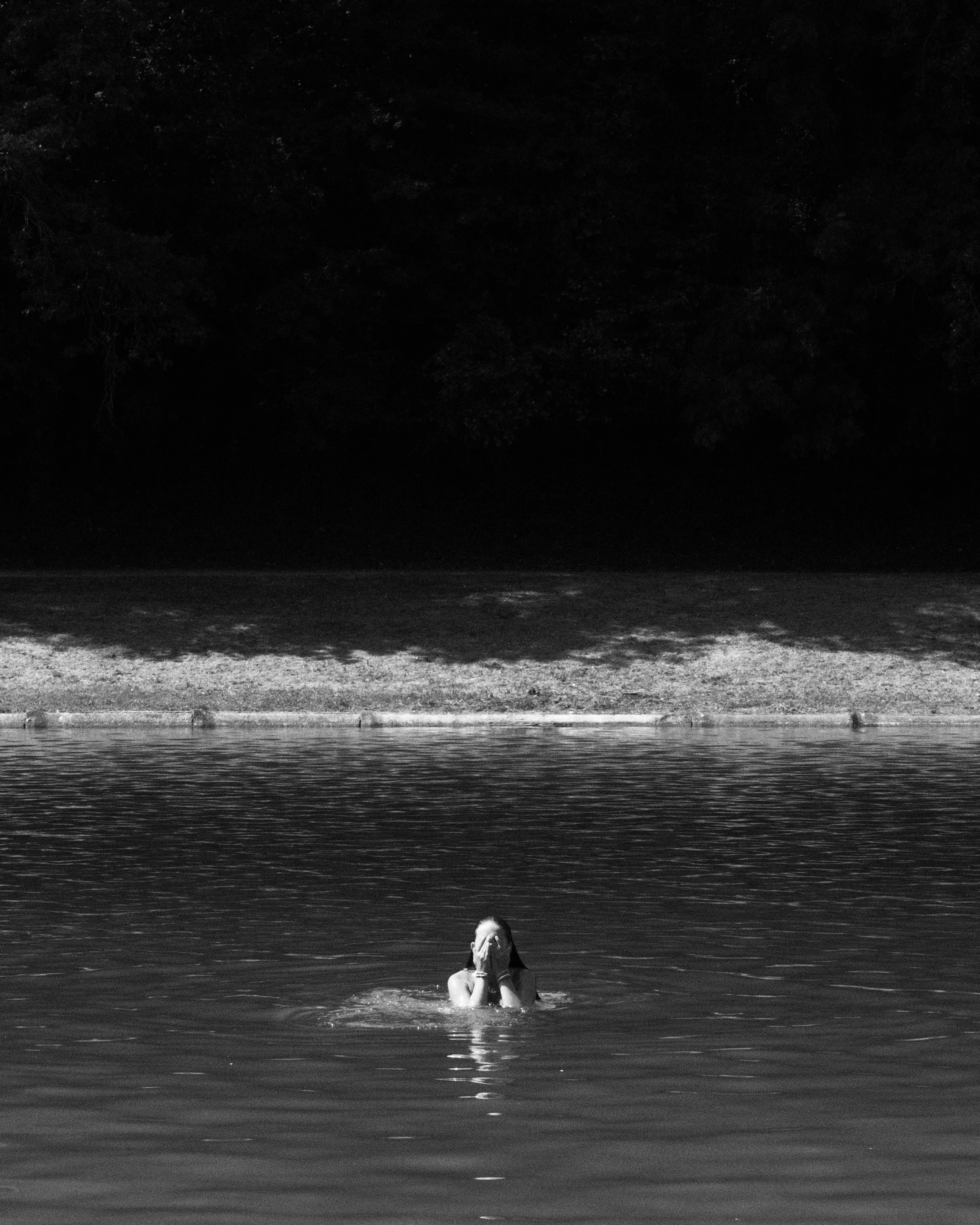 Une personne nage dans un lac, couvrant son visage avec ses mains, avec une forêt sombre en arrière-plan.