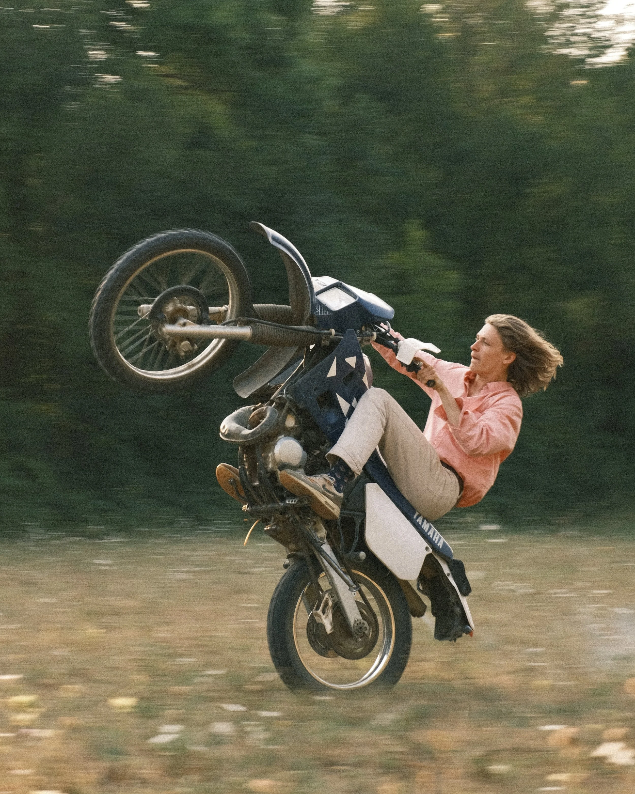 Une femme effectuant une roue arrière sur une moto Yamaha dans un champ avec des arbres en arrière-plan.