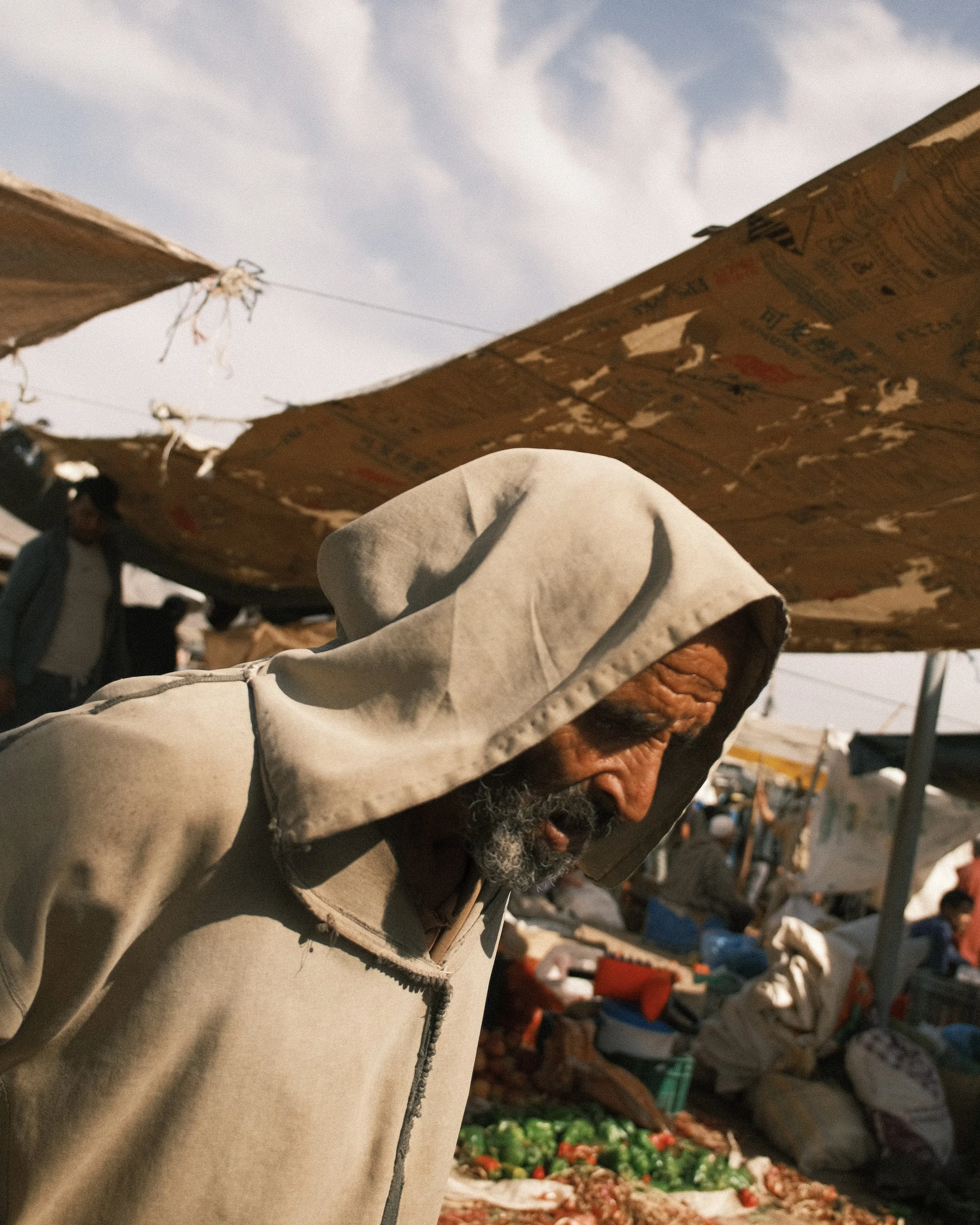 Un homme âgé portant une capuche beige à une foire ou un marché extérieur, avec des étals de légumes derrière lui et des tentes en toile au-dessus.