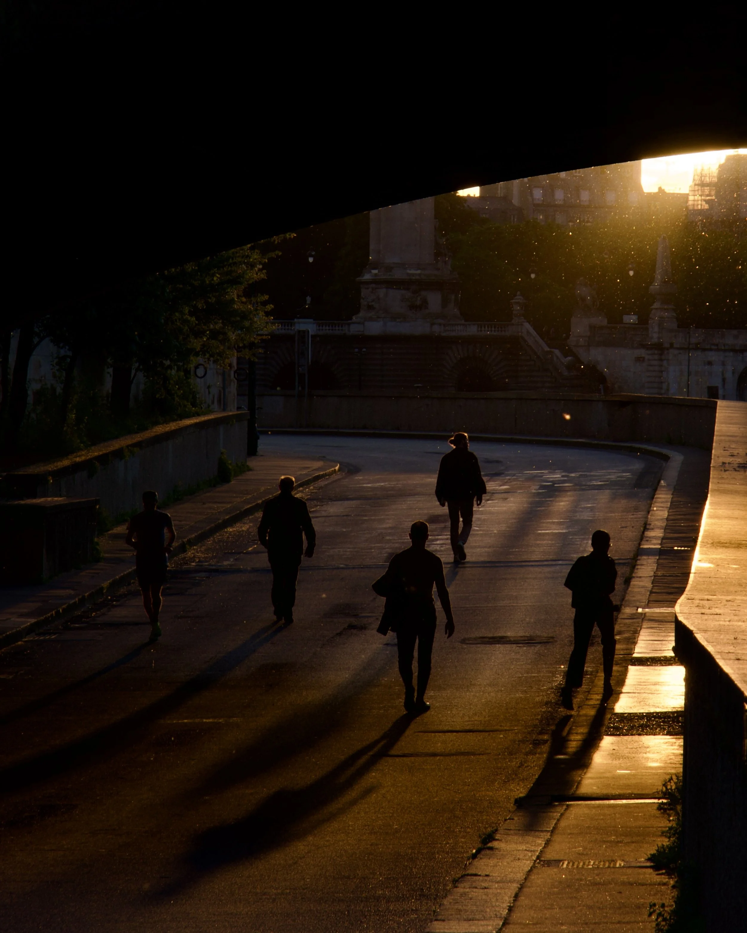 Groupe de personnes marchant sous un pont au coucher du soleil, avec des silhouettes noires et un horizon lumineux.