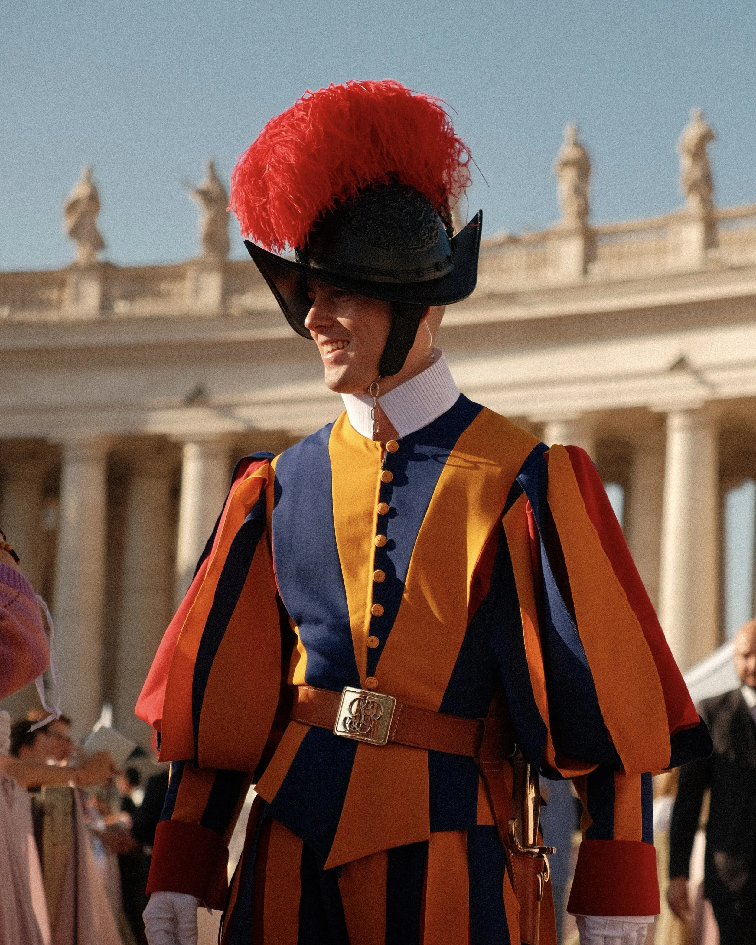 Un homme déguisé en membre de la garde nationale italienne, portant un uniforme coloré avec un casque à plumes rouges, en train de sourire lors d'une cérémonie en plein air devant un bâtiment historique avec des colonnes.