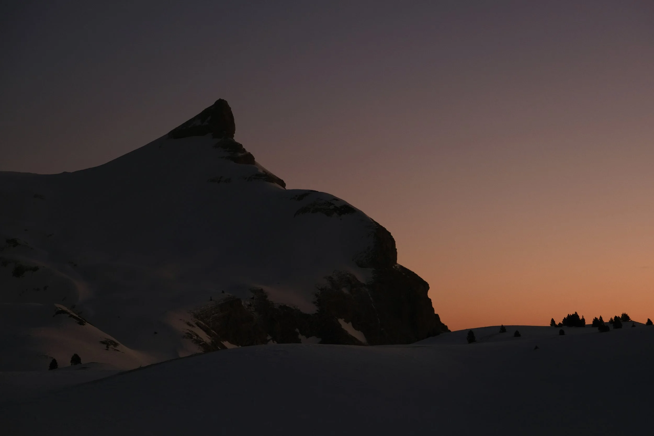 Montagne enneigée au coucher du soleil avec un ciel dégradé de couleurs chaudes et froides, silhouette de petits arbres en premier plan.