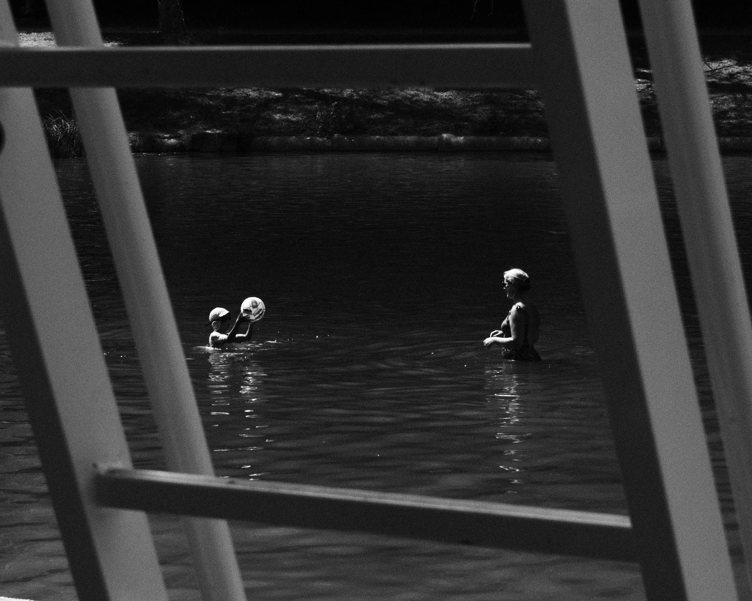 Une femme et un enfant jouent dans l'eau, vus entre les barreaux d'une chaise ou d'un banc en premier plan.