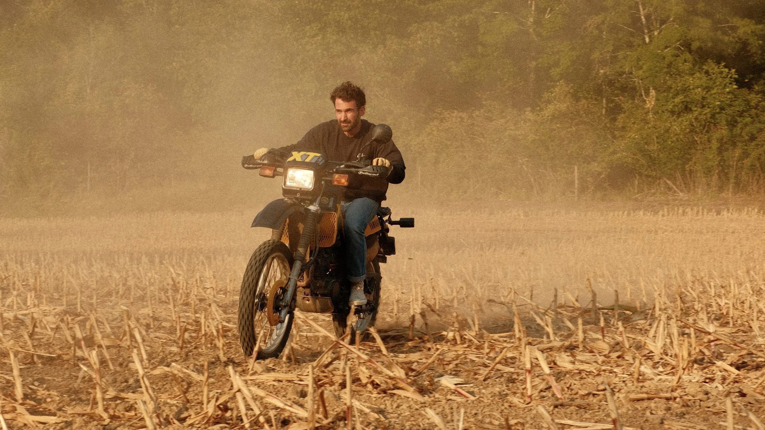 Un homme conduisant une moto dans un champ de maïs sec par une journée venteuse, avec des arbres en arrière-plan.