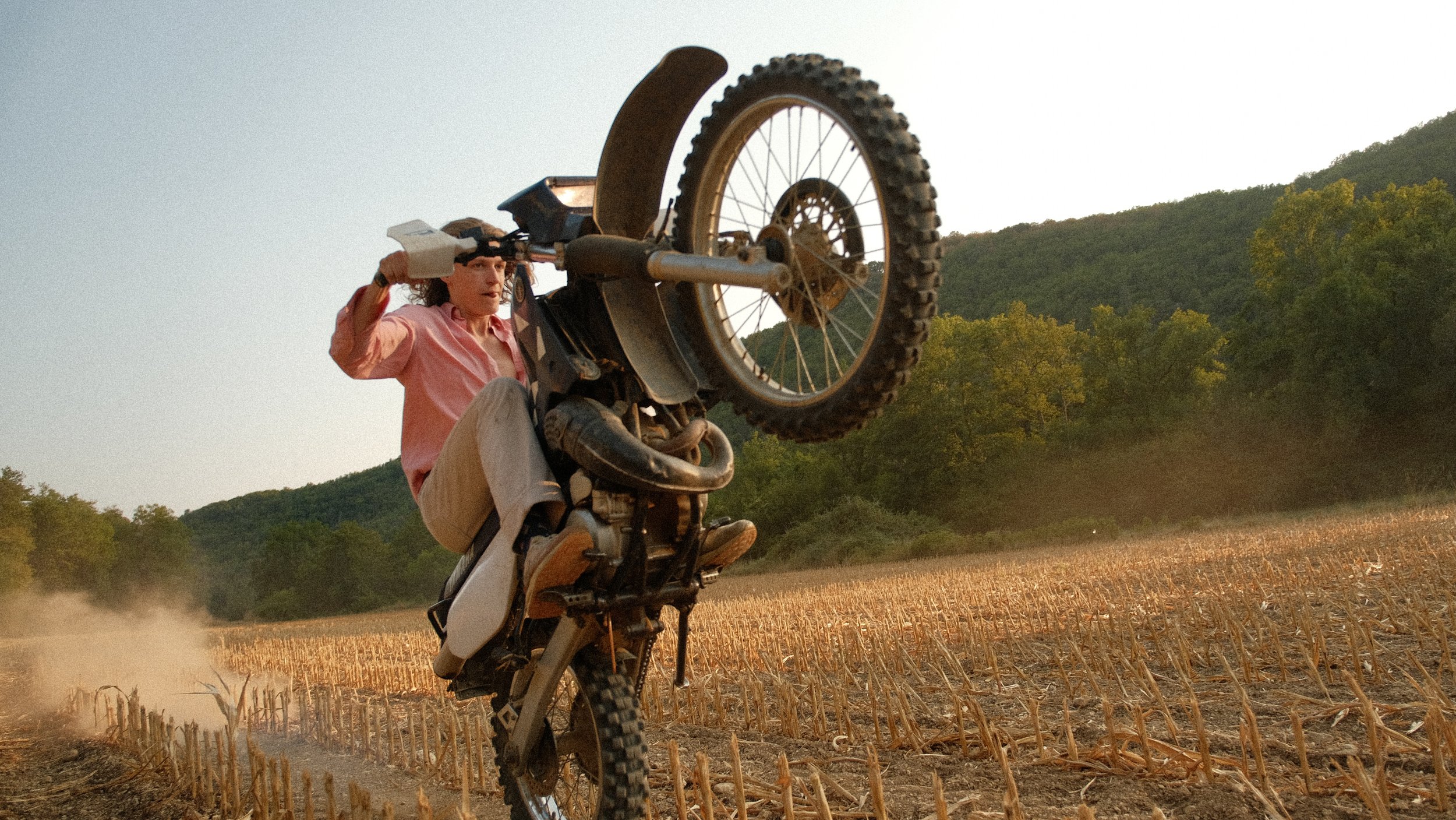 Une femme effectue une roue arrière sur une moto dans un champ, avec des montagnes en arrière-plan, lors du coucher de soleil.