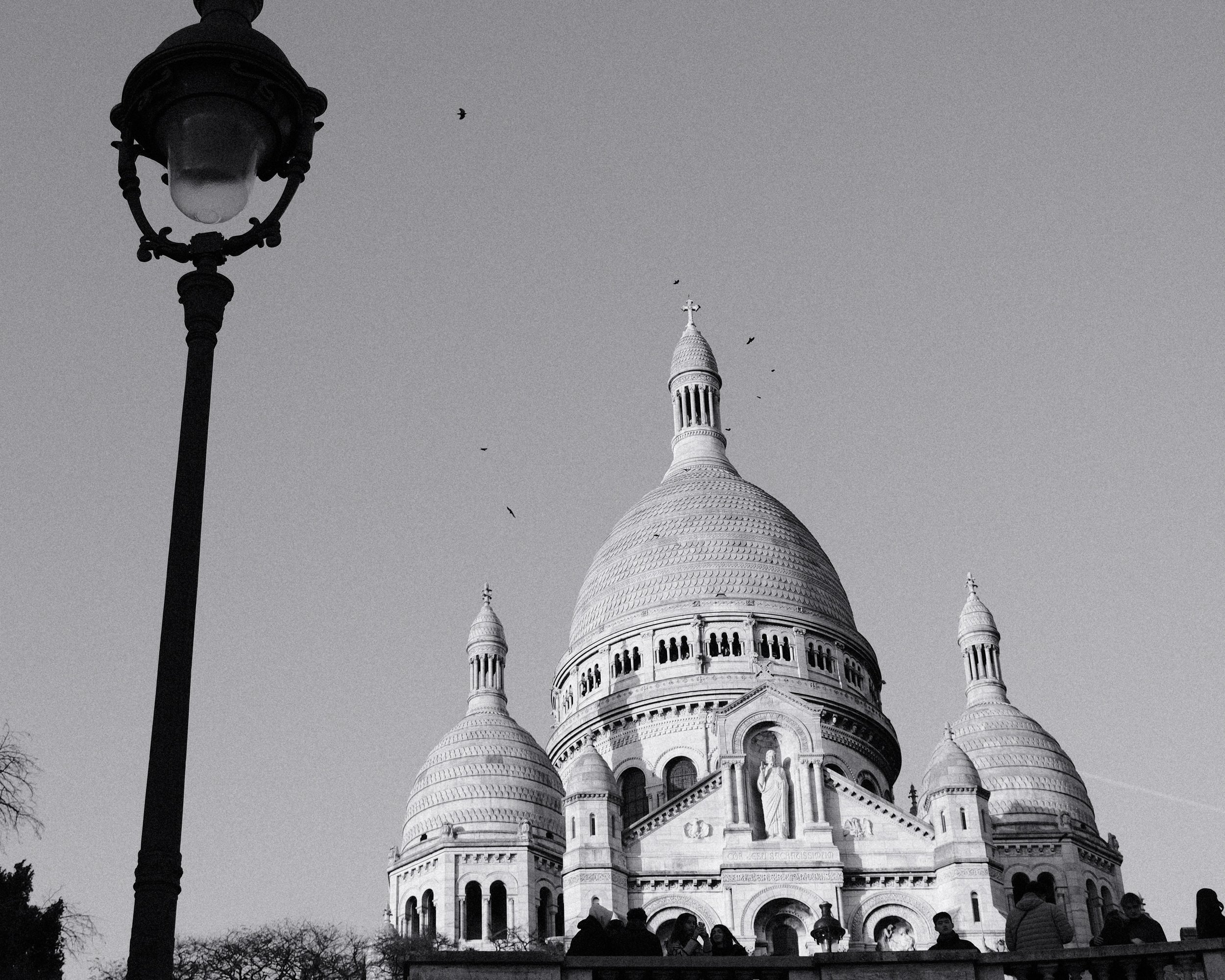 Photo en noir et blanc du Sacré-Cœur de Montmartre avec un lampadaire en premier plan, quelques oiseaux dans le ciel et des personnes en bas.