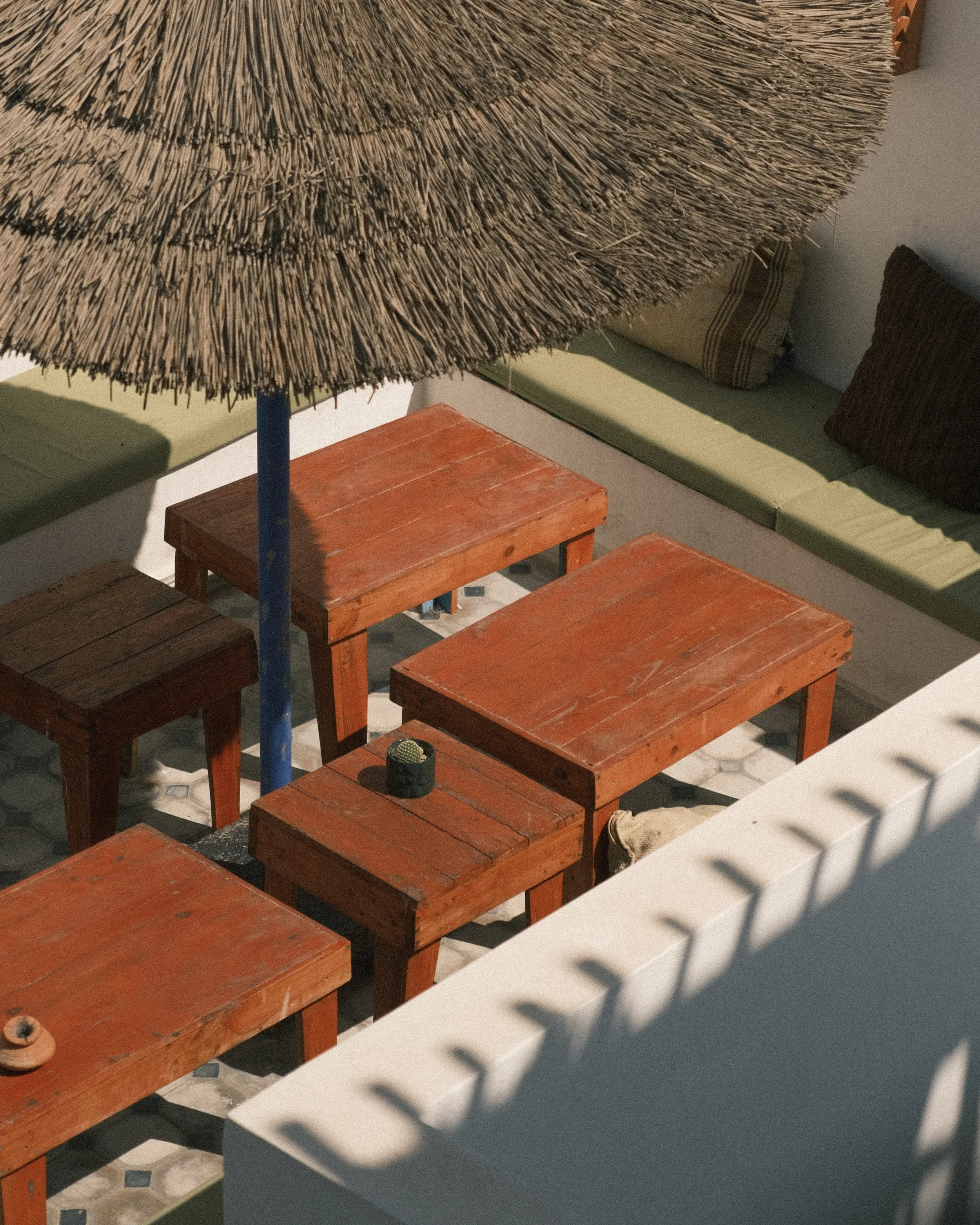 Vue d'une terrasse avec plusieurs tables en bois orange, un parasol en paille, et un banc avec coussins verts et marron.