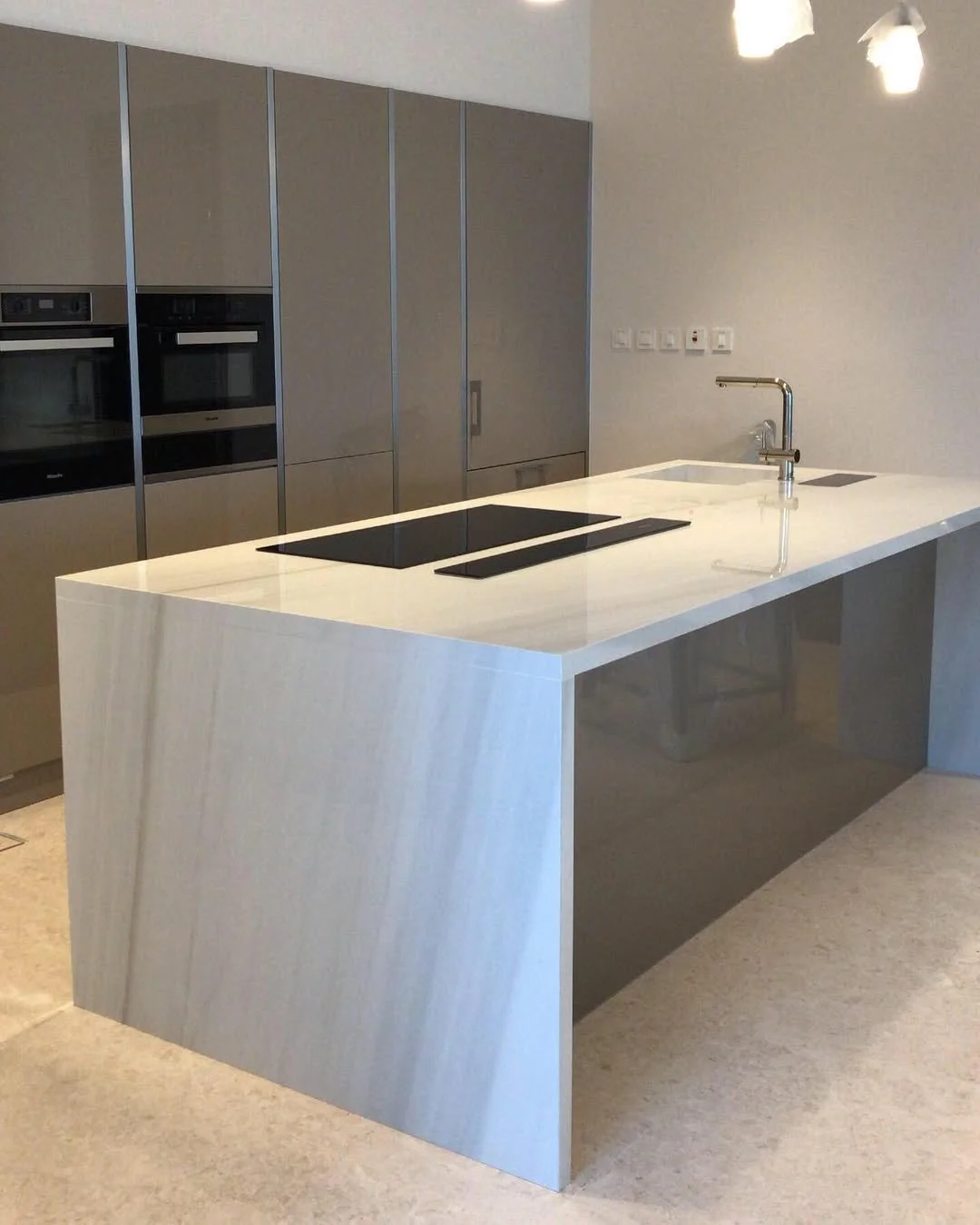 Modern kitchen with a sleek white island with a built-in black cooktop and a contemporary faucet, gray cabinets along the wall, and a wall with multiple electrical outlets.