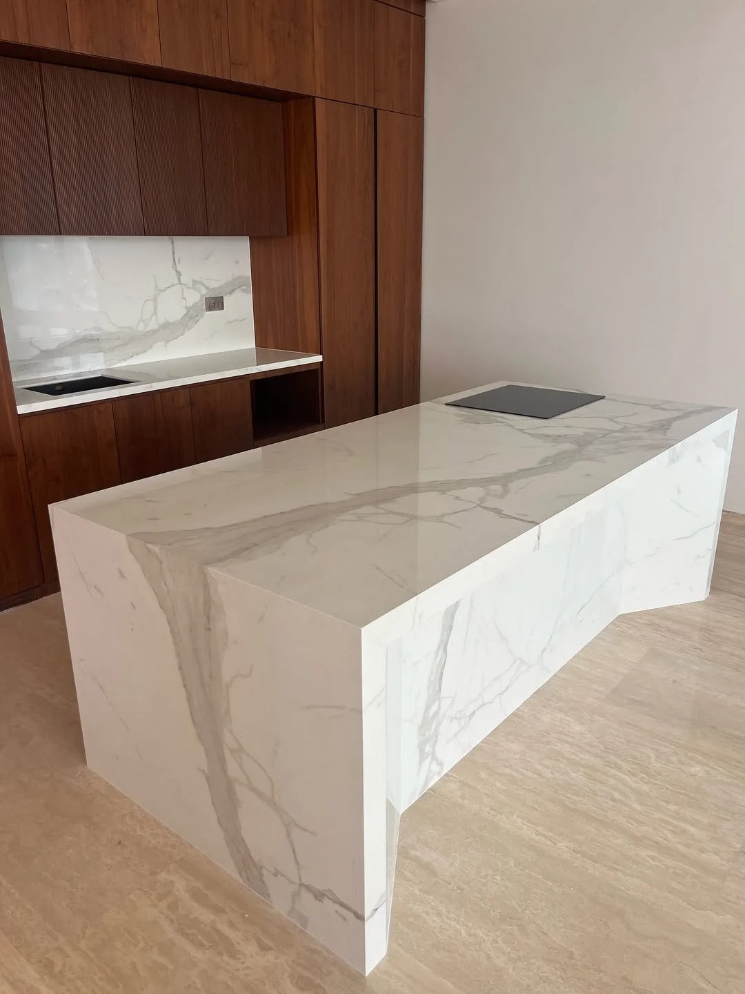 Modern kitchen with a white marble island with gray veining and a wooden wall cabinetry. A black stovetop is installed on the island, and there is a marble backsplash matching the island's material.