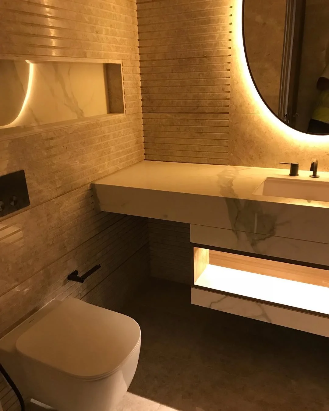 Modern bathroom with a white toilet, wooden wall panels, a white marble sink with dark fixtures, and a large circular mirror with backlighting.