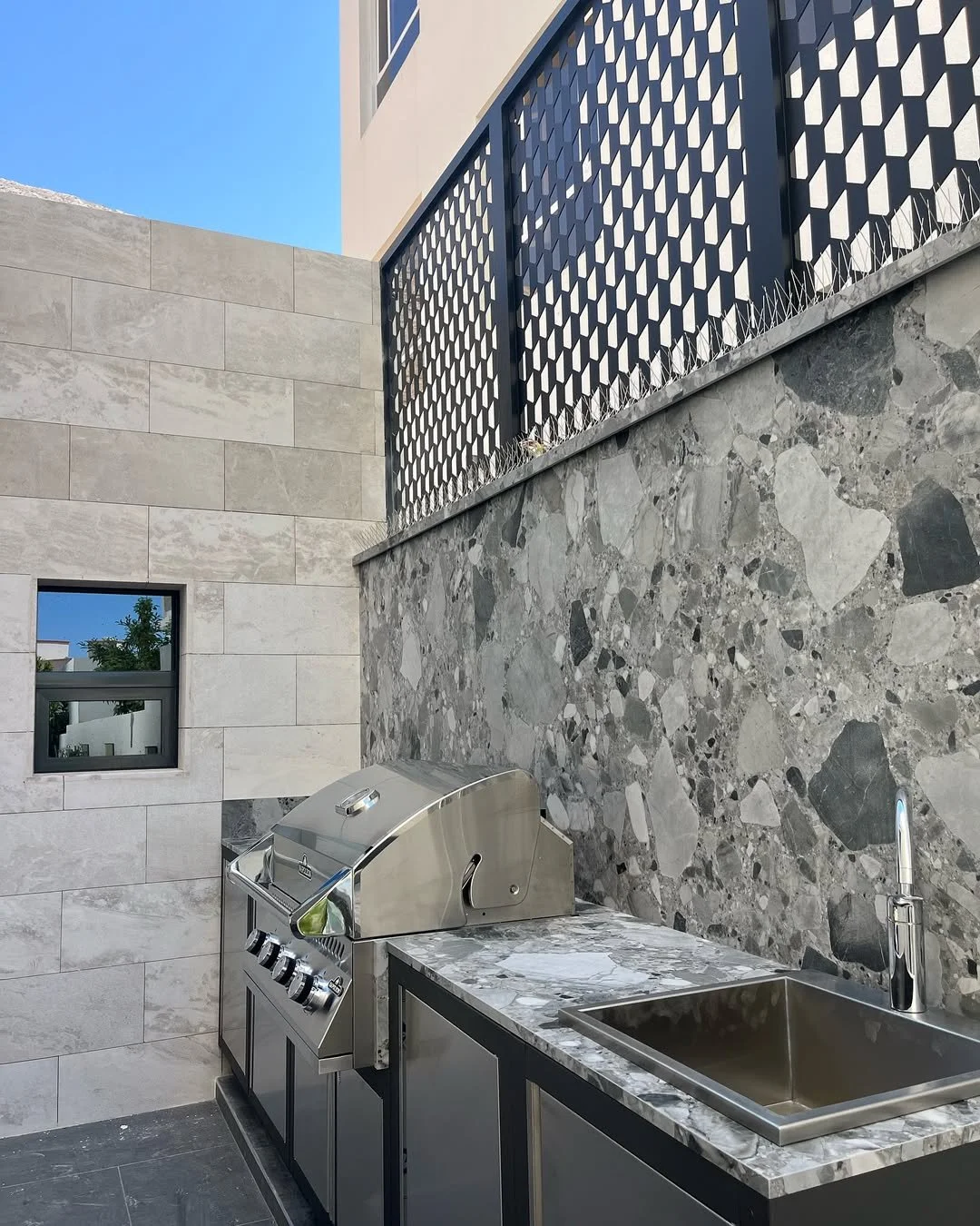 An outdoor kitchen area featuring a stainless steel grill, a marble countertop with a sink and modern faucet, and a stone wall with a privacy fence above.