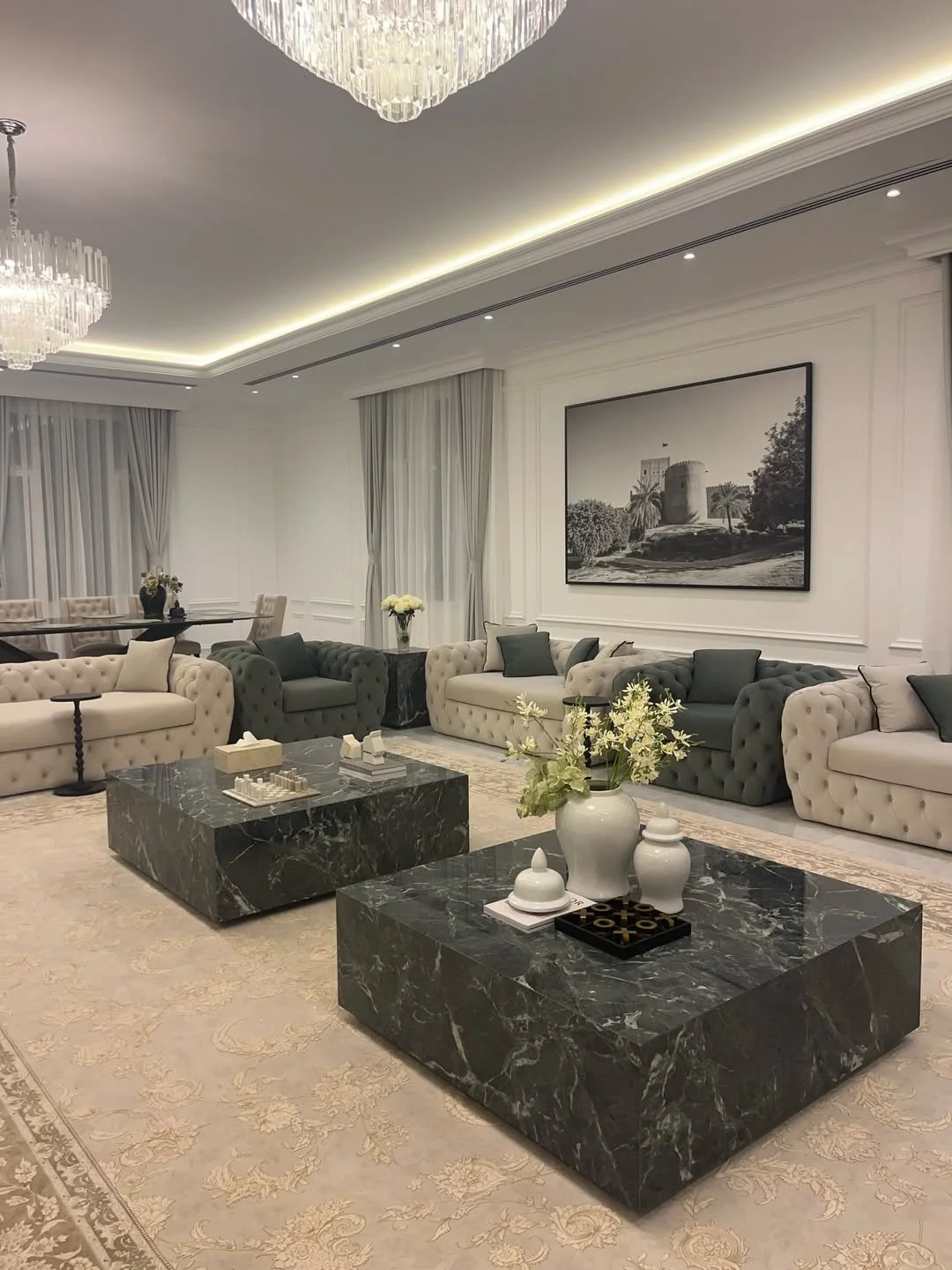 Elegant living room with white and green tufted sofas, marble coffee tables, floral arrangements, a black and white wall art, chandeliers, and large windows with curtains.