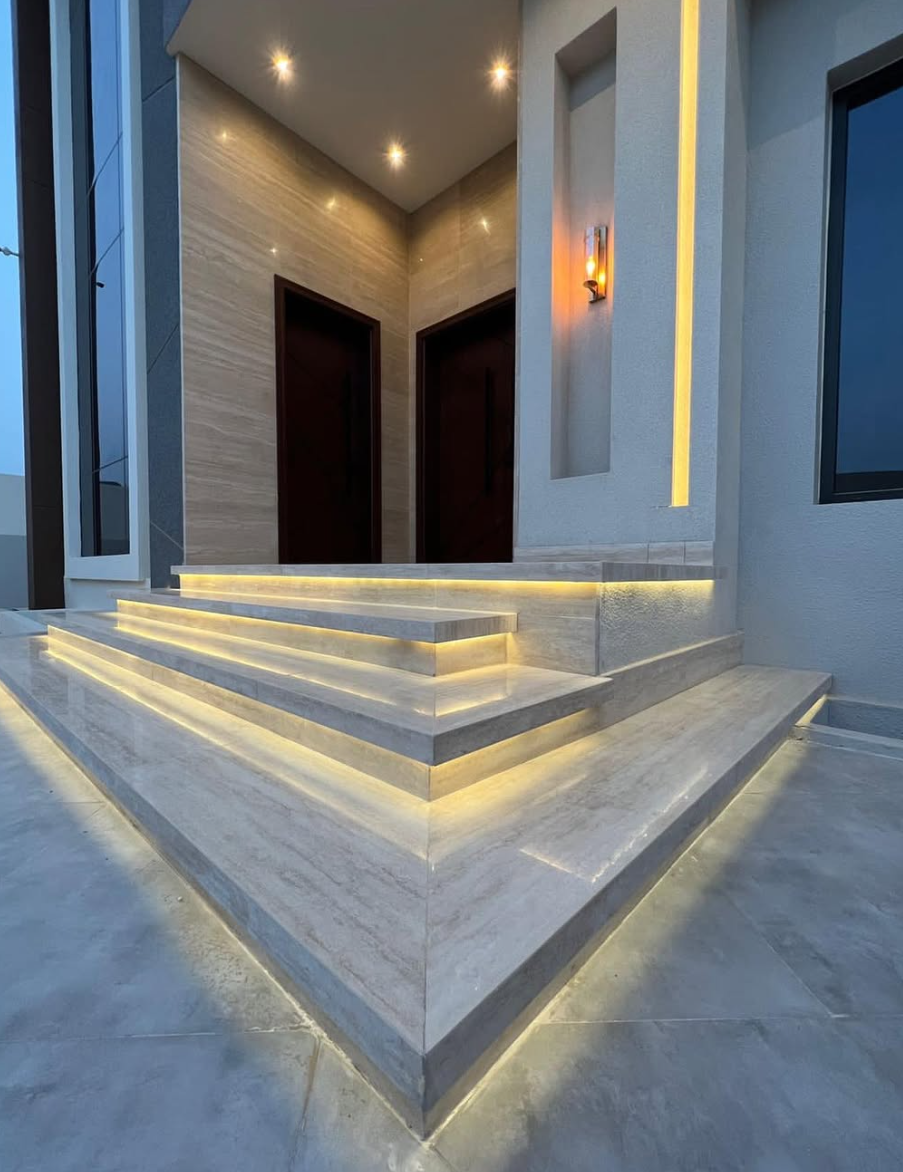 Modern house entrance with three marble steps, illuminated with yellow LED lighting, leading to a double door, with exterior wall lighting and a small niche with a light fixture.
