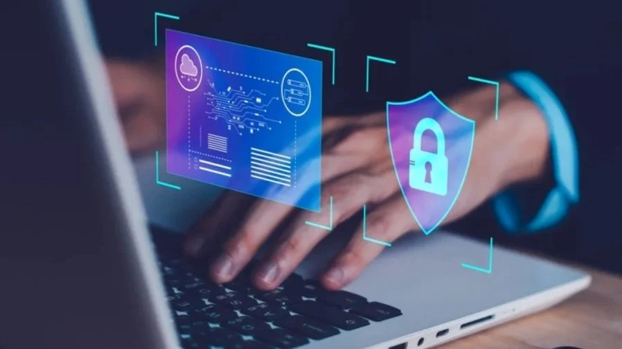 Person's hands using a laptop with digital security graphics overlay, including a cloud, circuit board, data, and a shield with a lock symbol, representing cybersecurity.