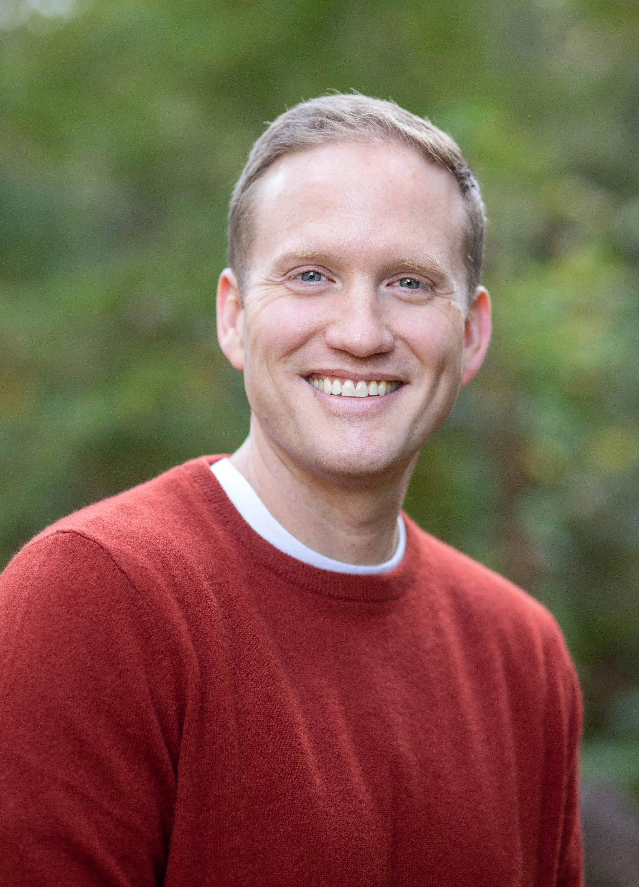 Portrait of a smiling man with blue eyes and short light brown hair, wearing a red sweater and a white shirt collar, outdoors with a blurred green background.