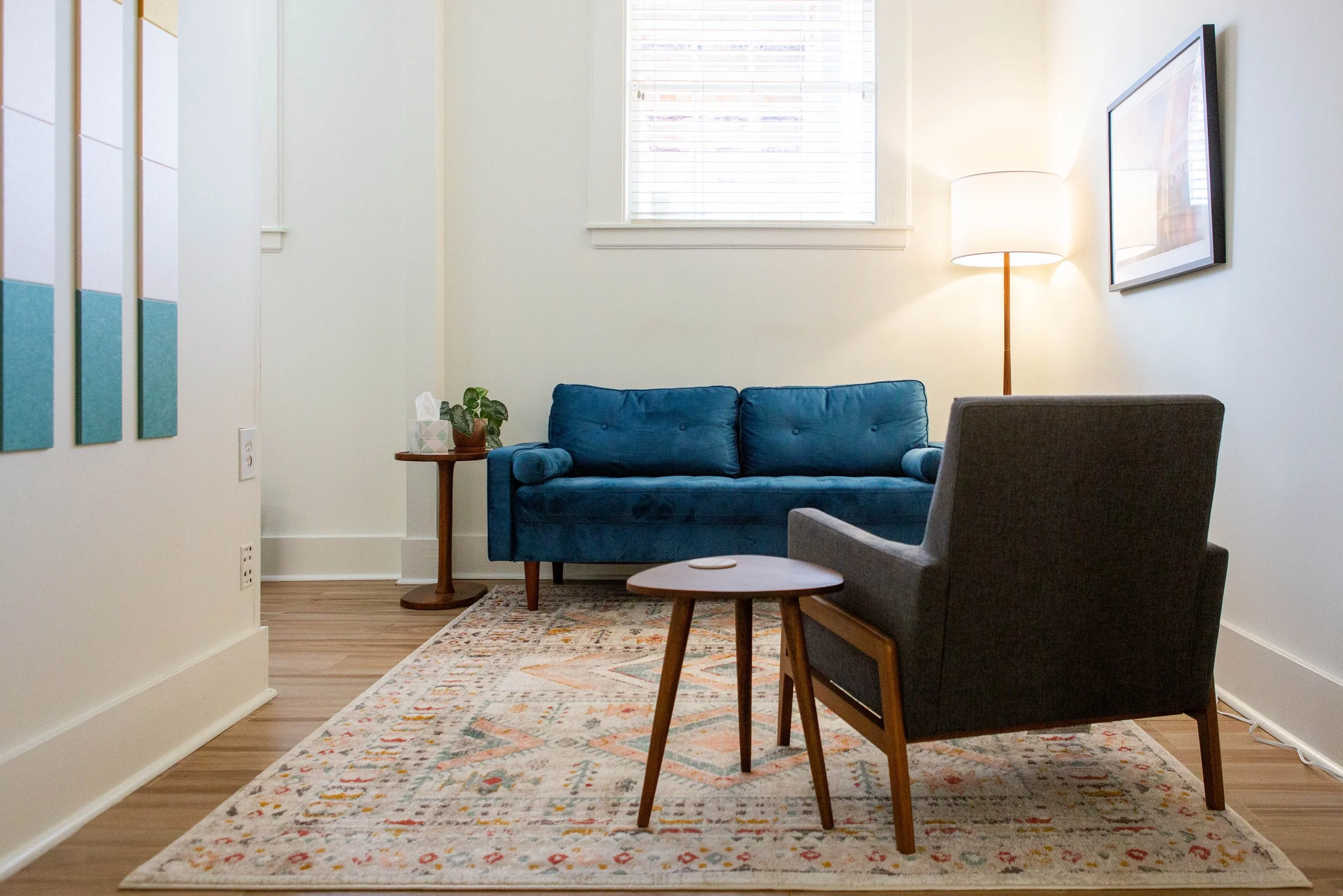 Cozy living room with a blue sofa, gray armchair, wooden side table with a plant, patterned rug, and a standing lamp near a window with blinds, decorated with framed art on the wall.