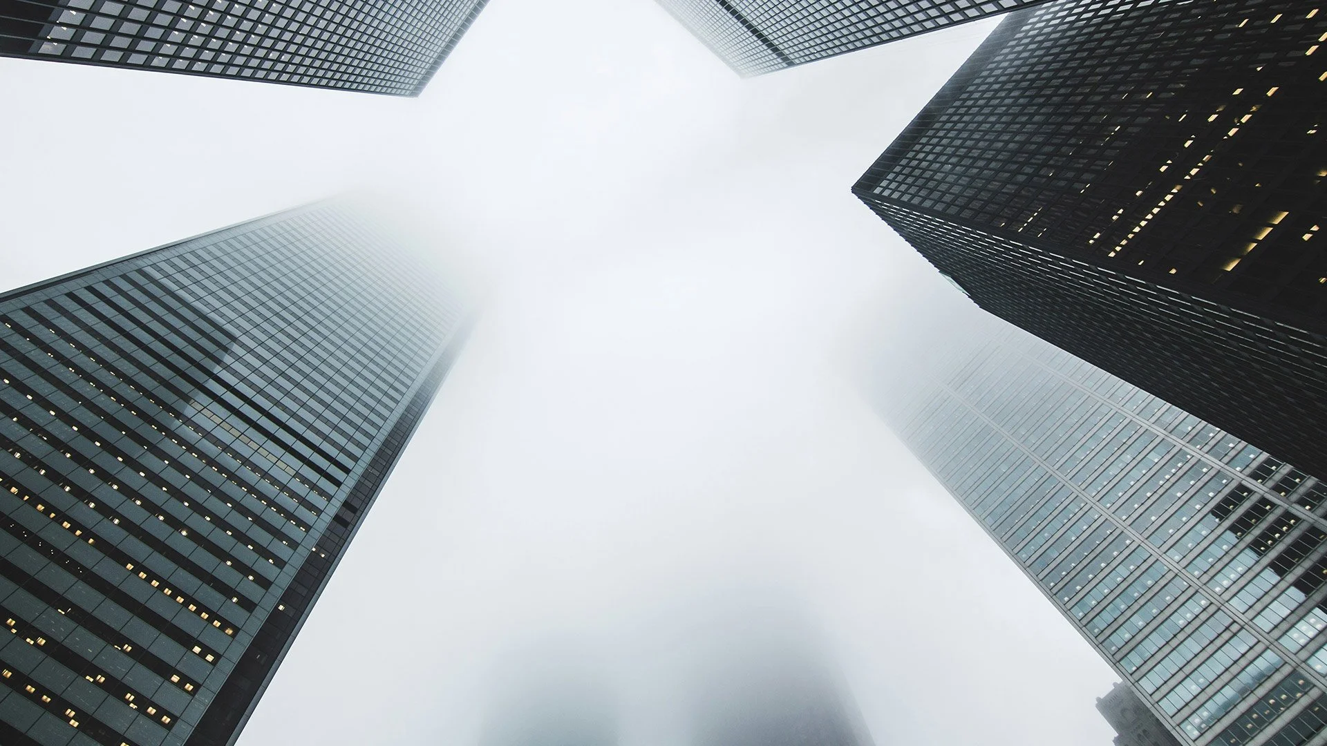 Looking up at tall modern skyscrapers in foggy weather with glass facades reflecting the surroundings.