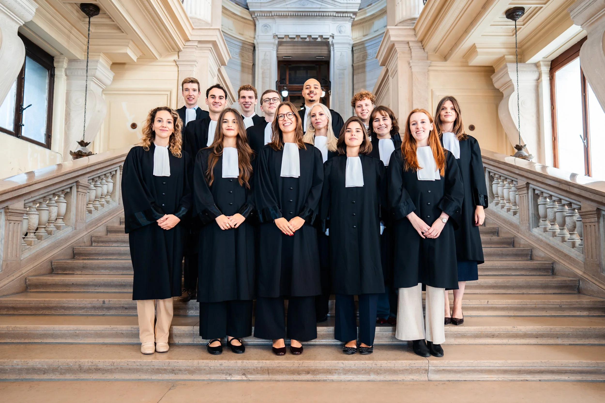 Groupe de jeunes adultes portant des robes de graduation noirs, posant sur un escalier en marbre dans une salle élégante avec des détails architecturaux classiques.