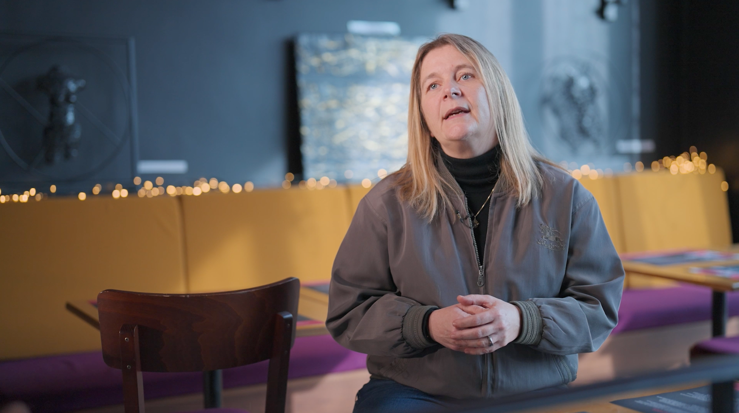 Une femme parle dans un restaurant ou un café, assise à une table avec des chaises en arrière-plan, décorée avec des lumières chaudes et des œuvres d'art sur les murs.