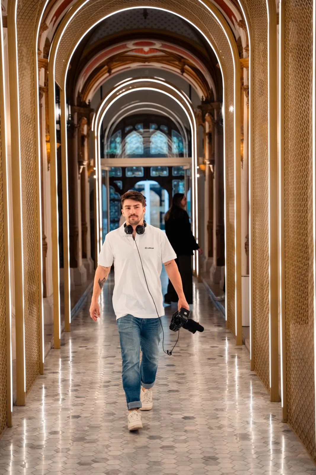 A man walking through a decorated, illuminated hallway with a camera in his hand and headphones around his neck.
