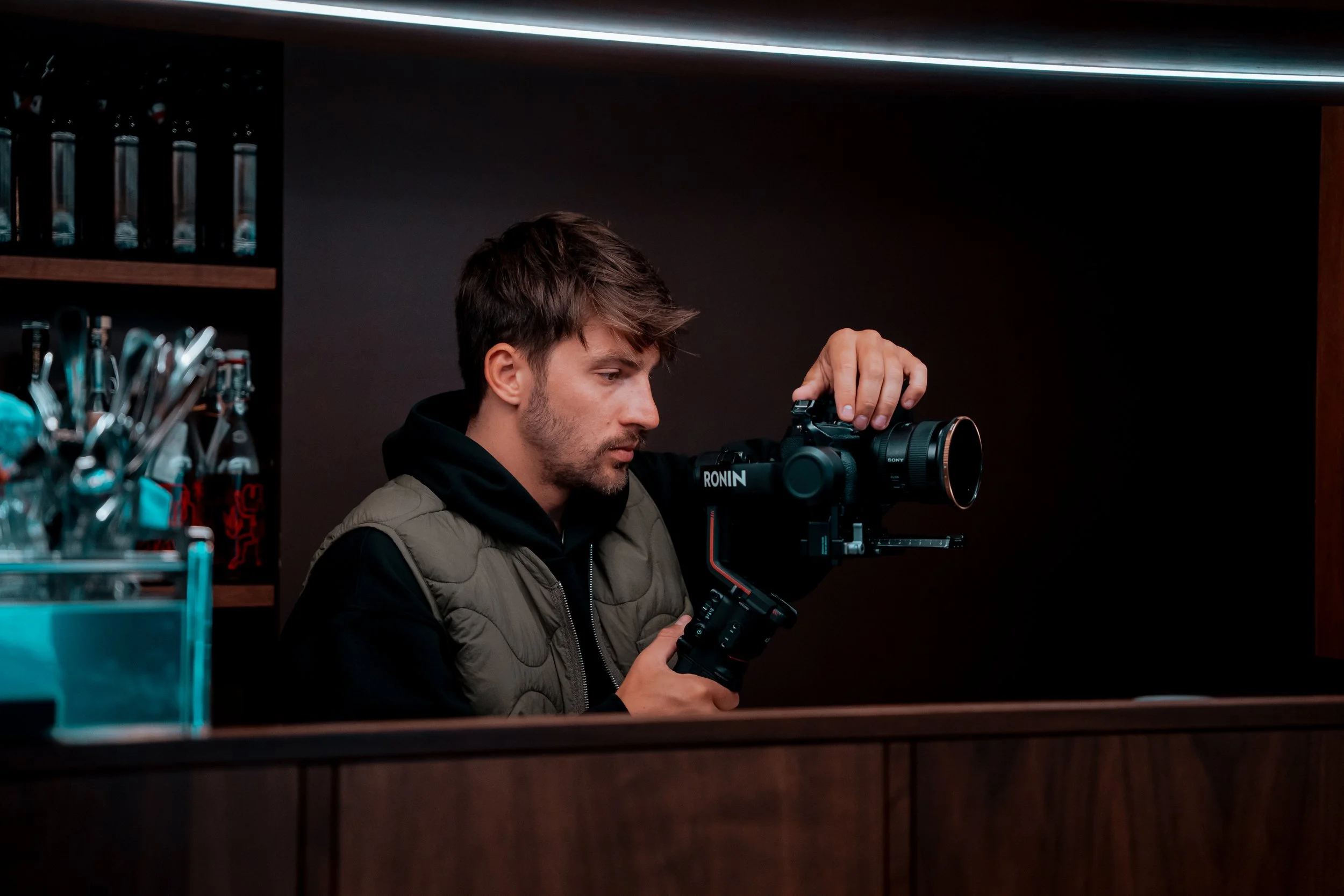 A man with brown hair and a beard operating a camera on a tripod in a dimly lit room.