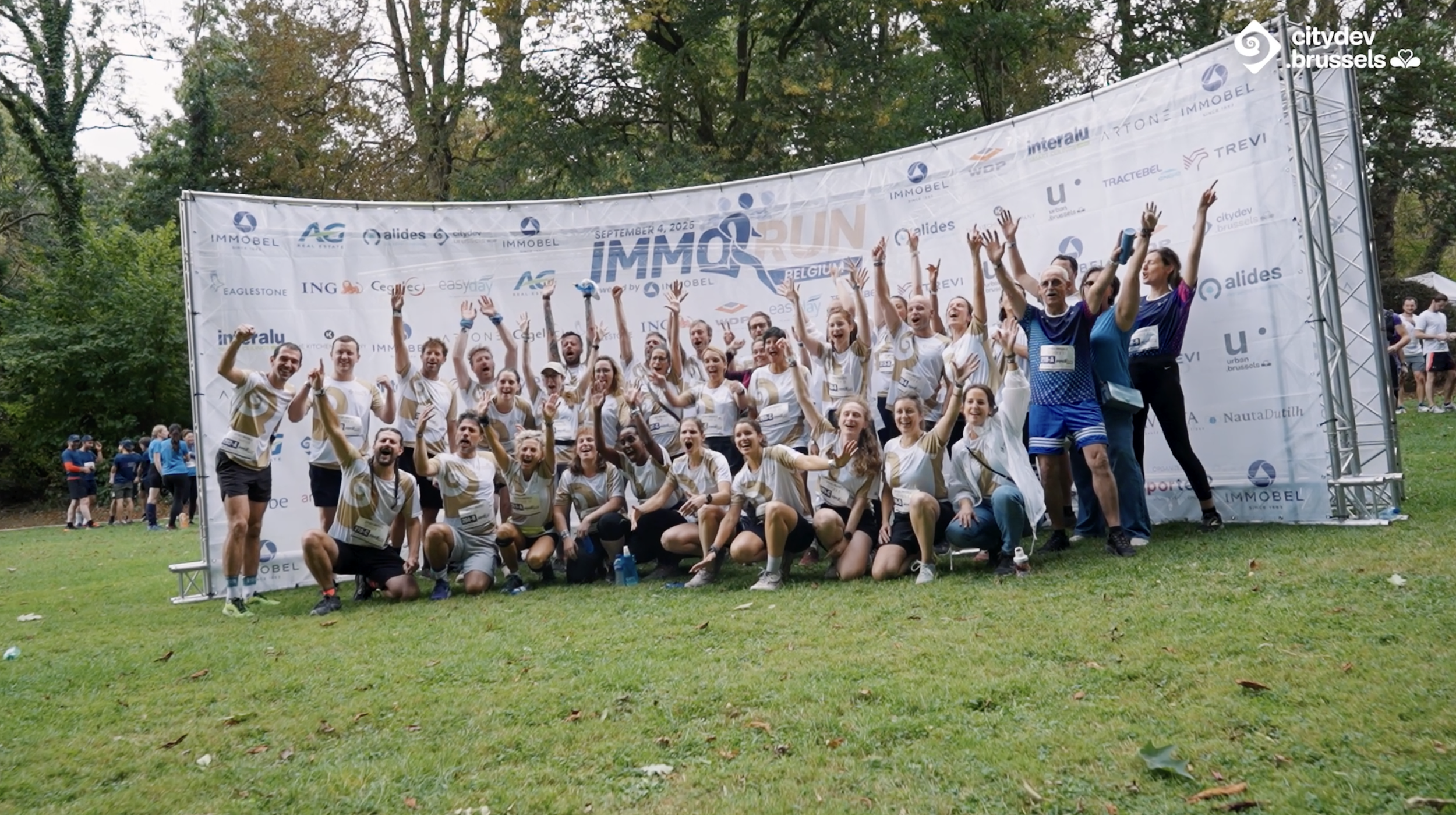 Groupe de coureurs et bénévoles posant souriants devant un panneau de l'événement IMMORUN, au parc, en pleine matinée.