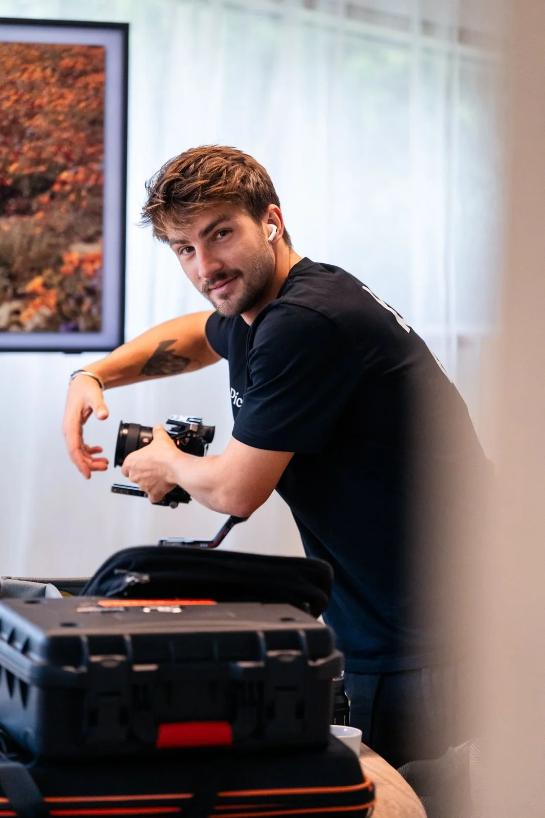 A young man with brown hair and a beard is holding camera equipment at a table, looking at the camera, with a print of autumn leaves on a screen in the background.