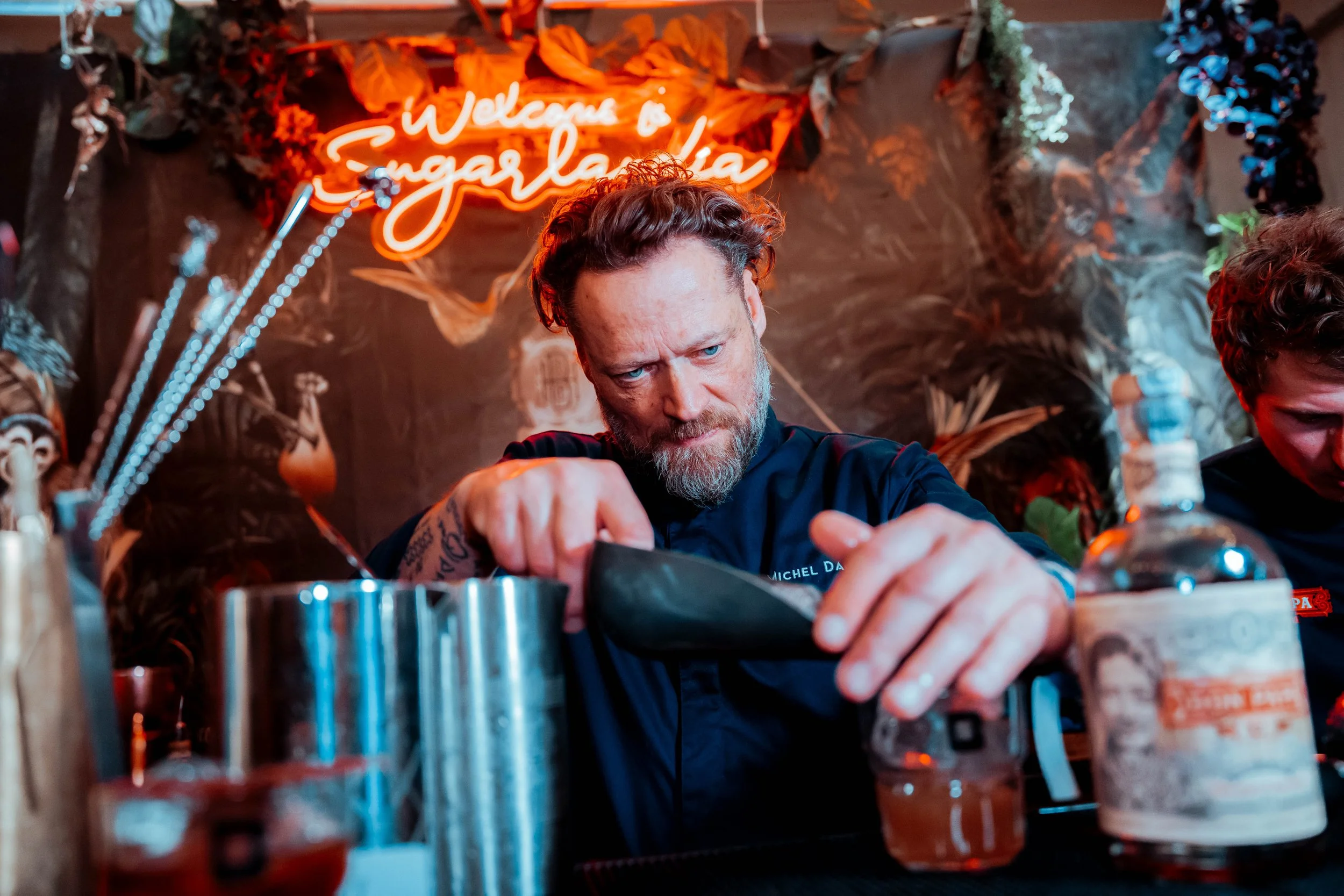 Un homme avec une barbe et des cheveux bouclés travaille dans un bar ou un restaurant, devant un comptoir avec diverses bouteilles et accessoires. En arrière-plan, un panneau lumineux en néon écrit 'Welcome to EngaVita'.