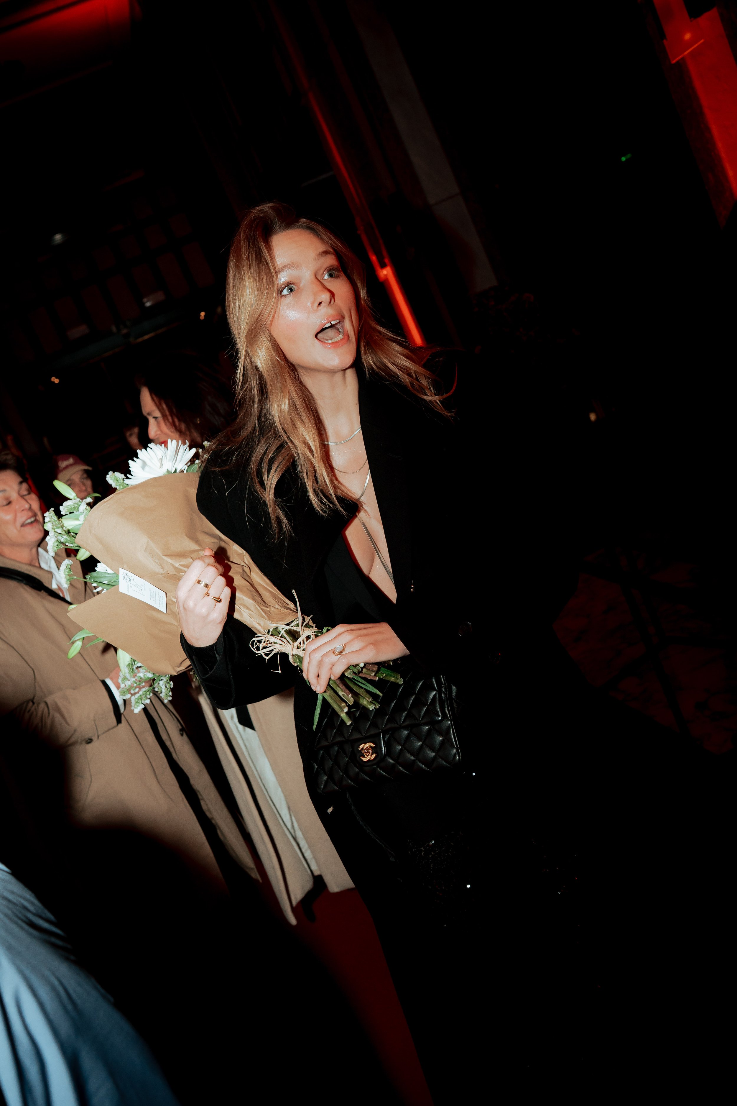 A woman with long blonde hair holding a bouquet of flowers and a black purse, smiling and looking surprised.