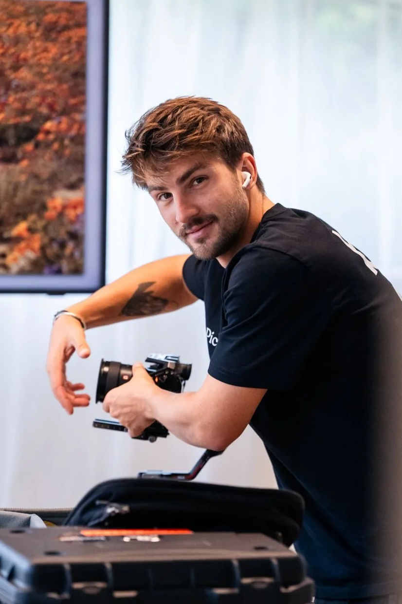 Jeune homme avec tatouage et écouteurs, tenant un stabilisateur pour caméra, dans un intérieur lumineux.