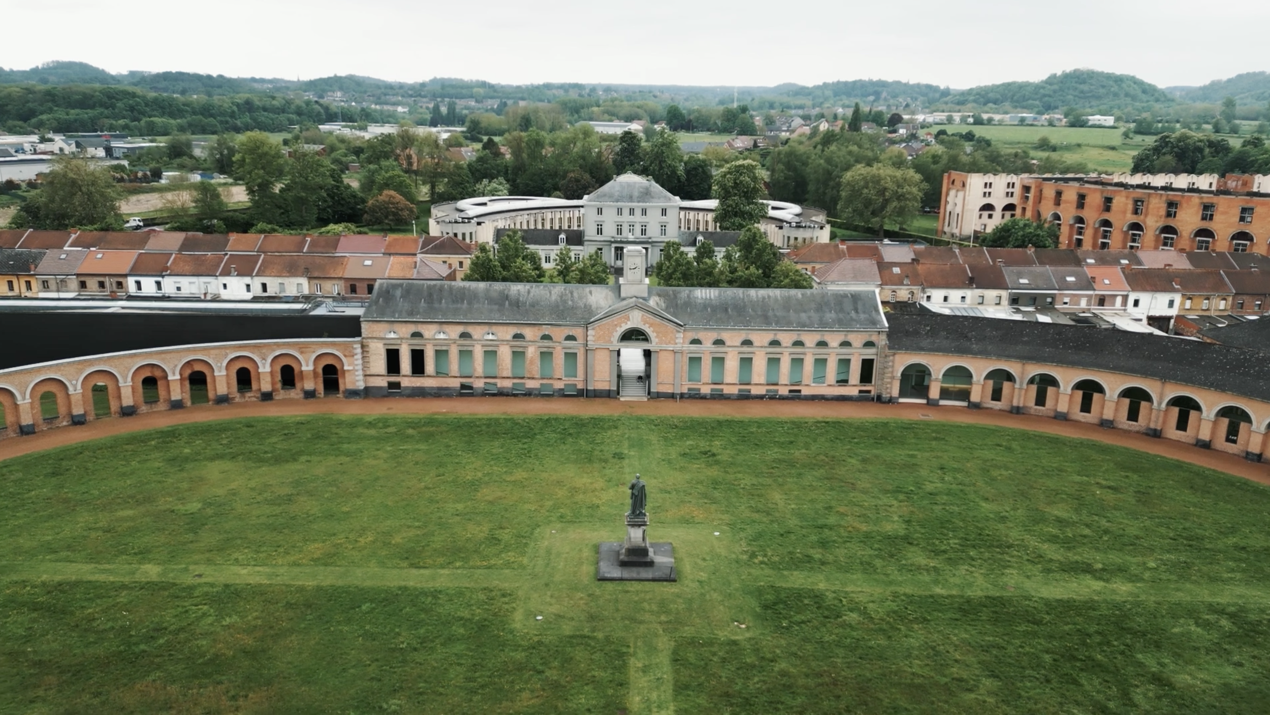 Vue aérienne d'un bâtiment historique avec un jardin en avant et un monument au centre, entouré de toits en tuiles et d'une végétation abondante.