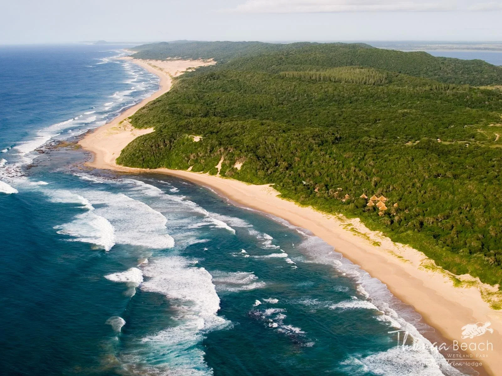 Aerial view of a sandy beach along a coastline with waves crashing onto the shore, green forested hills behind, and a few thatched-roof huts visible among the trees.