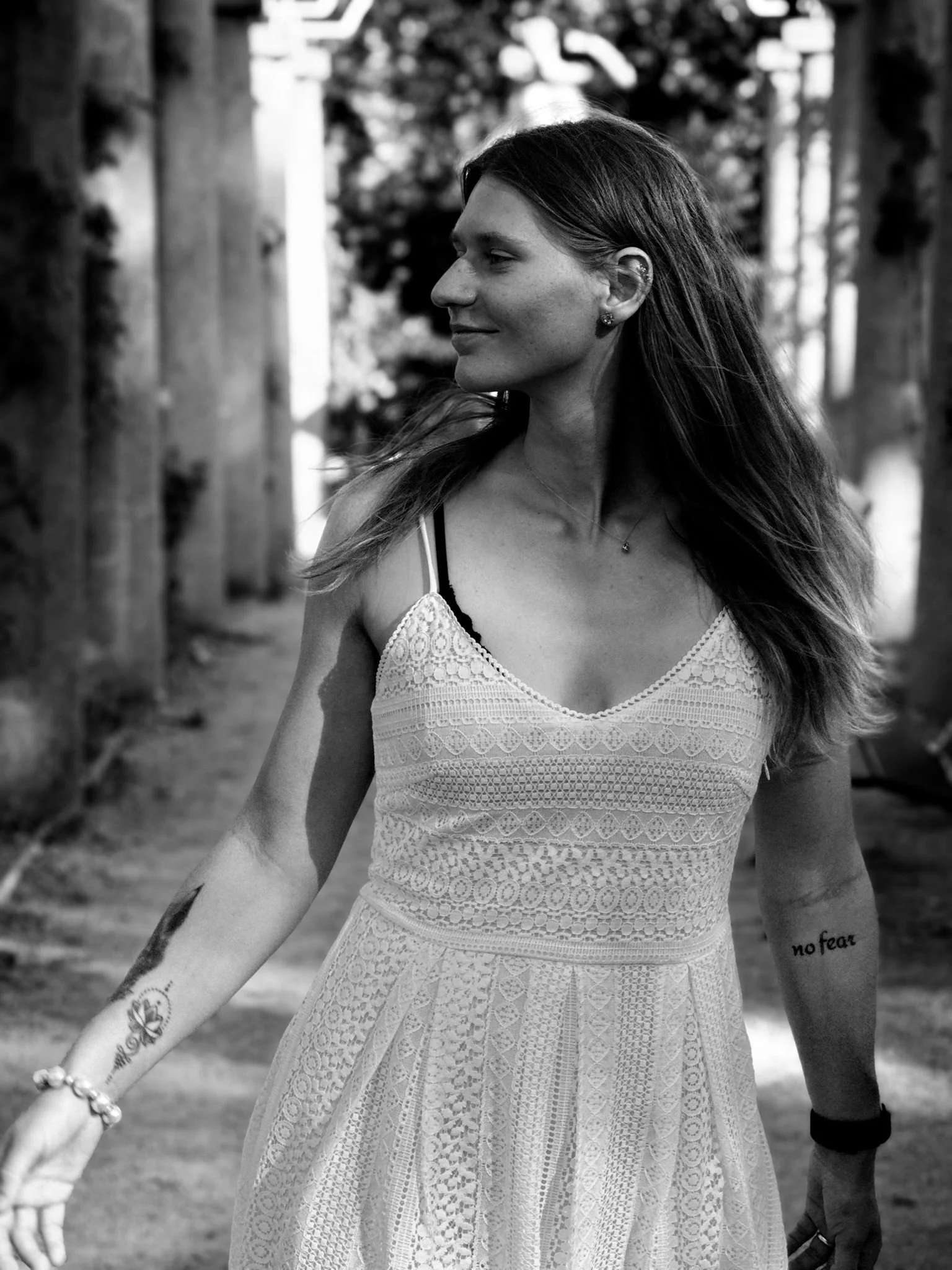 Eine junge Frau mit langen Haaren, die ein stilvolles, ärmelloses, weißes, geblümtes Kleid trägt, steht im Freien zwischen Bäumen in einer Holzpassage und blickt seitwärts.