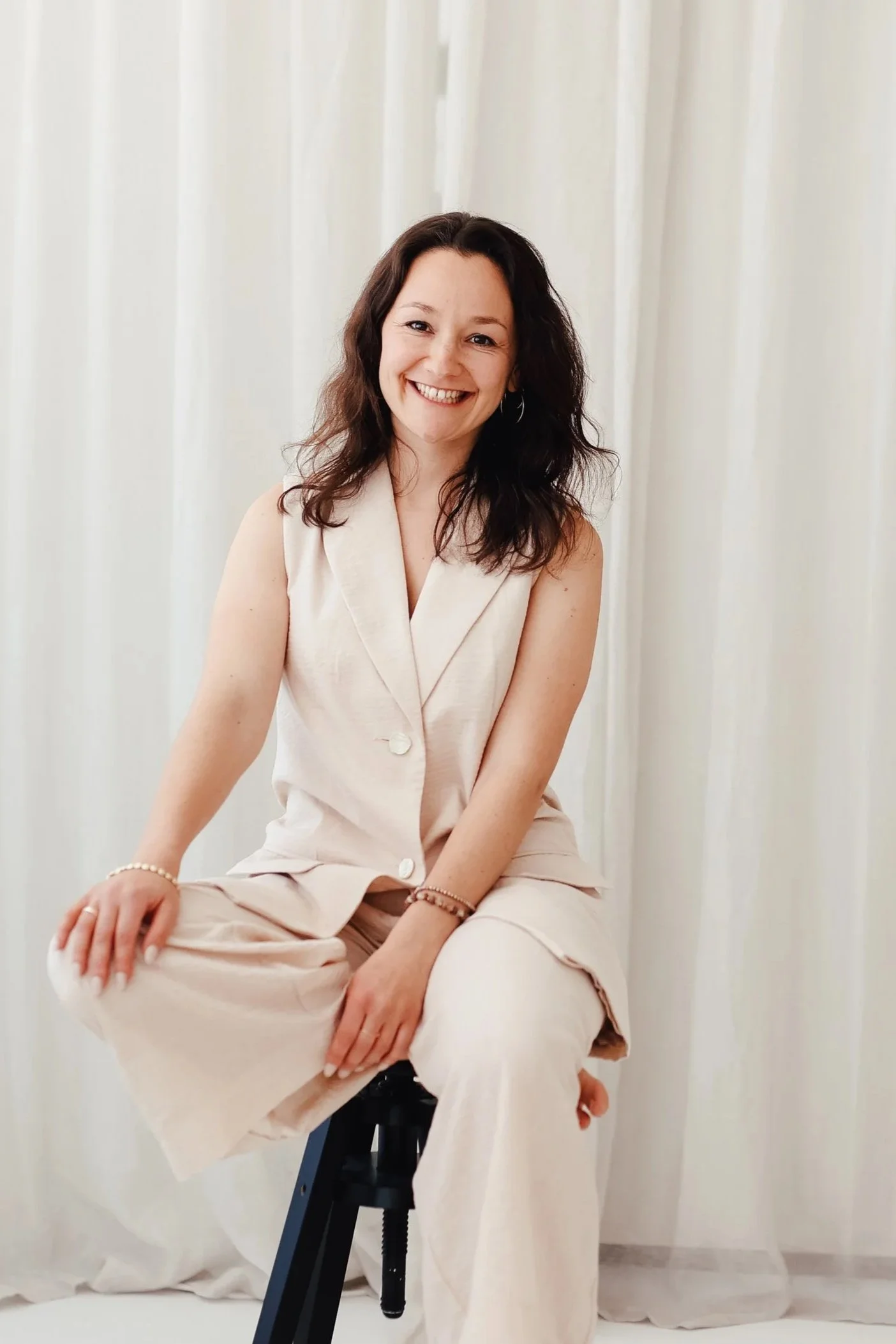 Felicia Thiering, systemische Beraterin & Elterncoach mit dunklen, lockigen Haaren in einem hellen, ärmellosen Anzug sitzt auf einem Hocker vor einem hellen Vorhang.