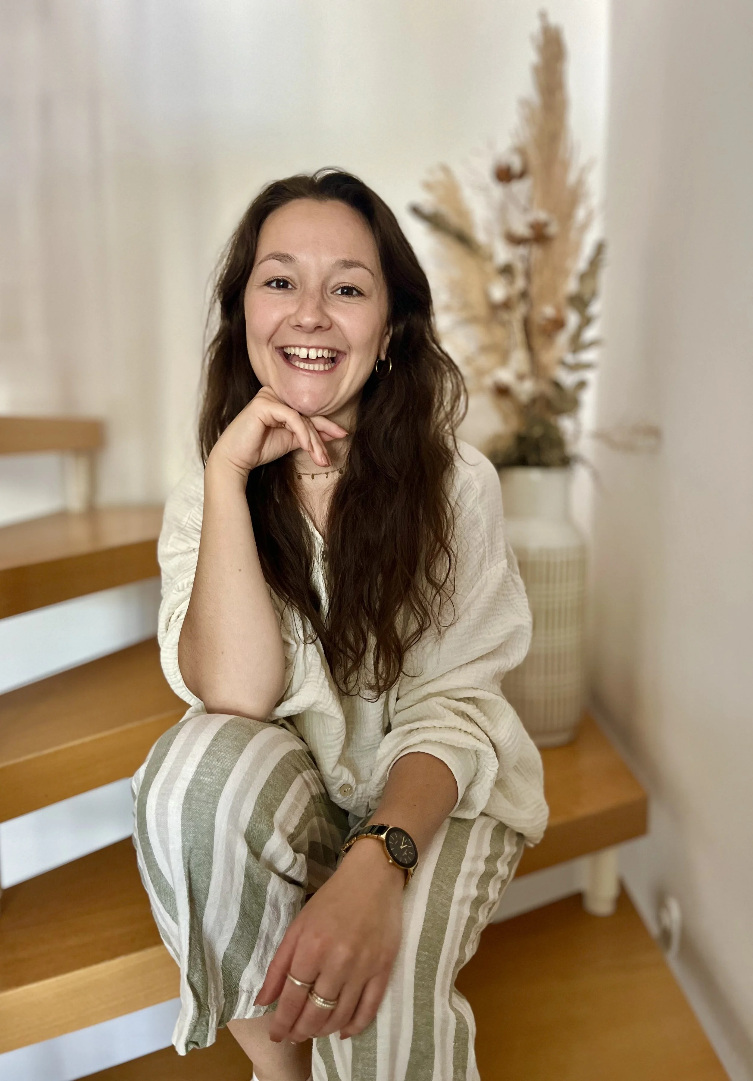 Ein lächelnder junger Frau sitzt auf einer Treppe in einem Wohnzimmer, trägt gestreifte Hosen und eine hellfarbene Bluse, trägt eine Armbanduhr, mehrere Ringe und einen dünnen Halsreifen, hat lange braune Haare, und im Hintergrund steht eine dekorative Pflanze.