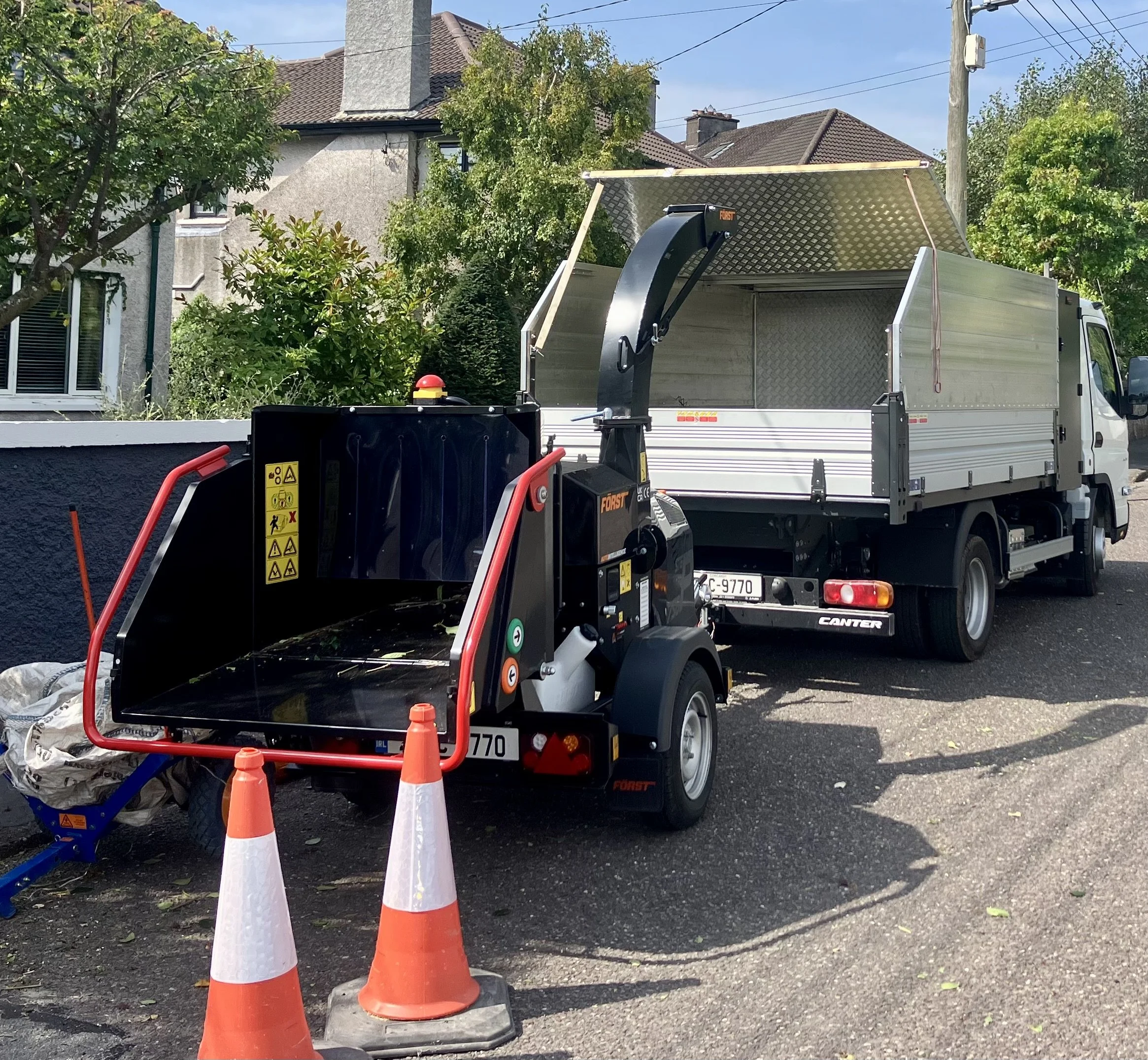 A street scene with a street sweeper machine and a white truck parked nearby, orange traffic cones in front, residential houses and trees in the background.