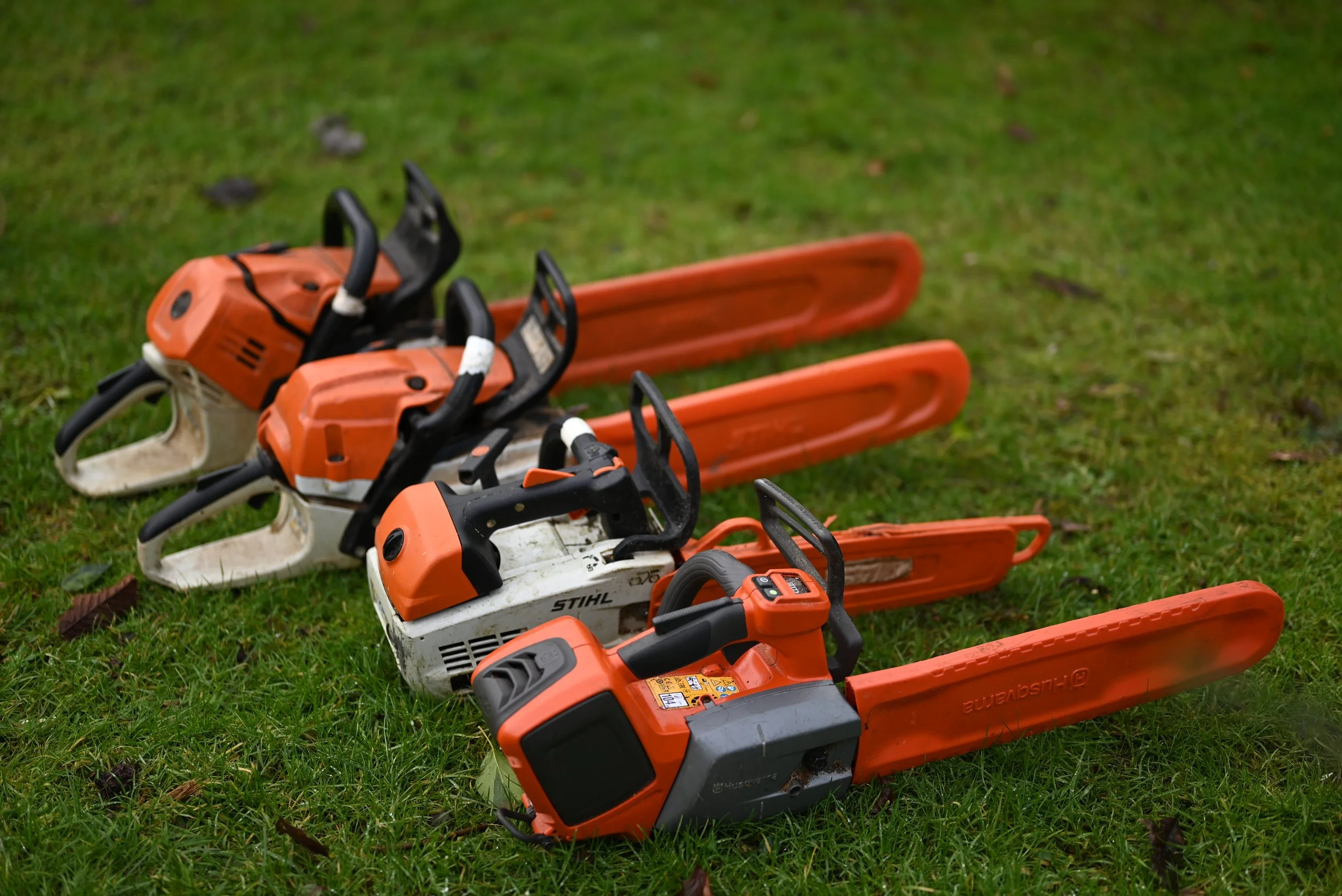 Three chainsaws with orange blades and black handles lying on green grass.