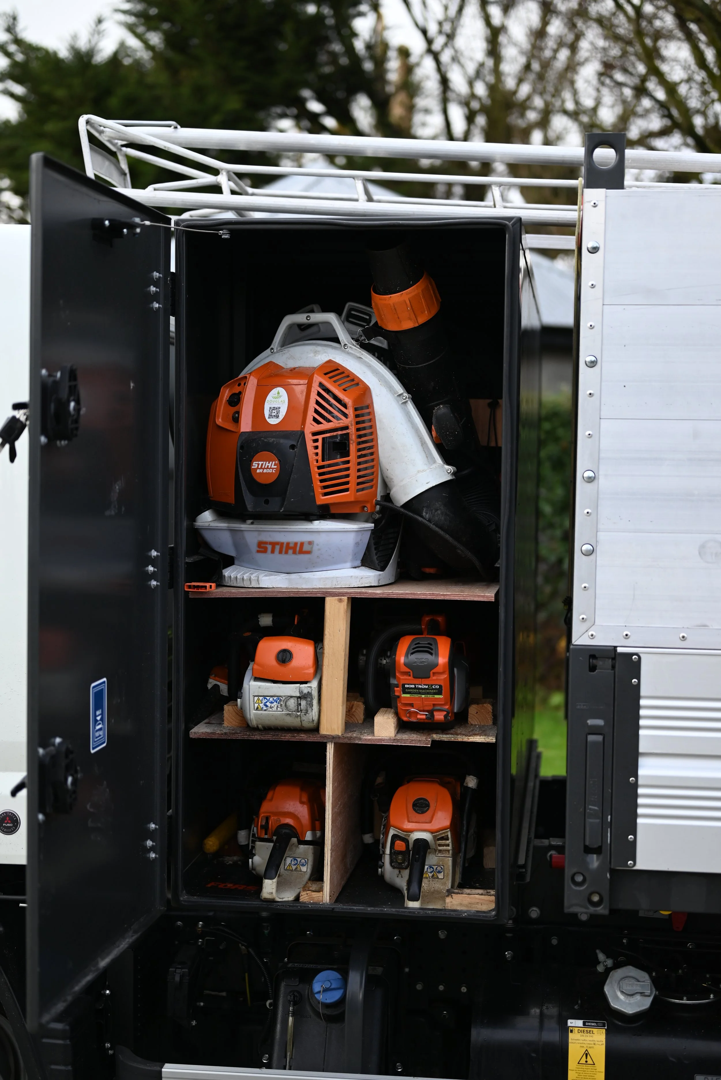Open storage compartment on a service truck containing Stihl brand outdoor power tools, including a leaf blower and chainsaws, organized on wooden shelves.