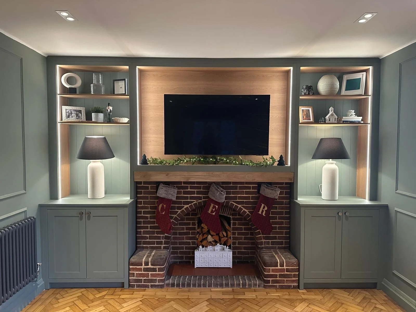 Living room fireplace with a mounted flat-screen TV, surrounded by built-in shelves with decorative items, lamps on both sides, and Christmas stockings hanging on the brick fireplace with firewood stored underneath.