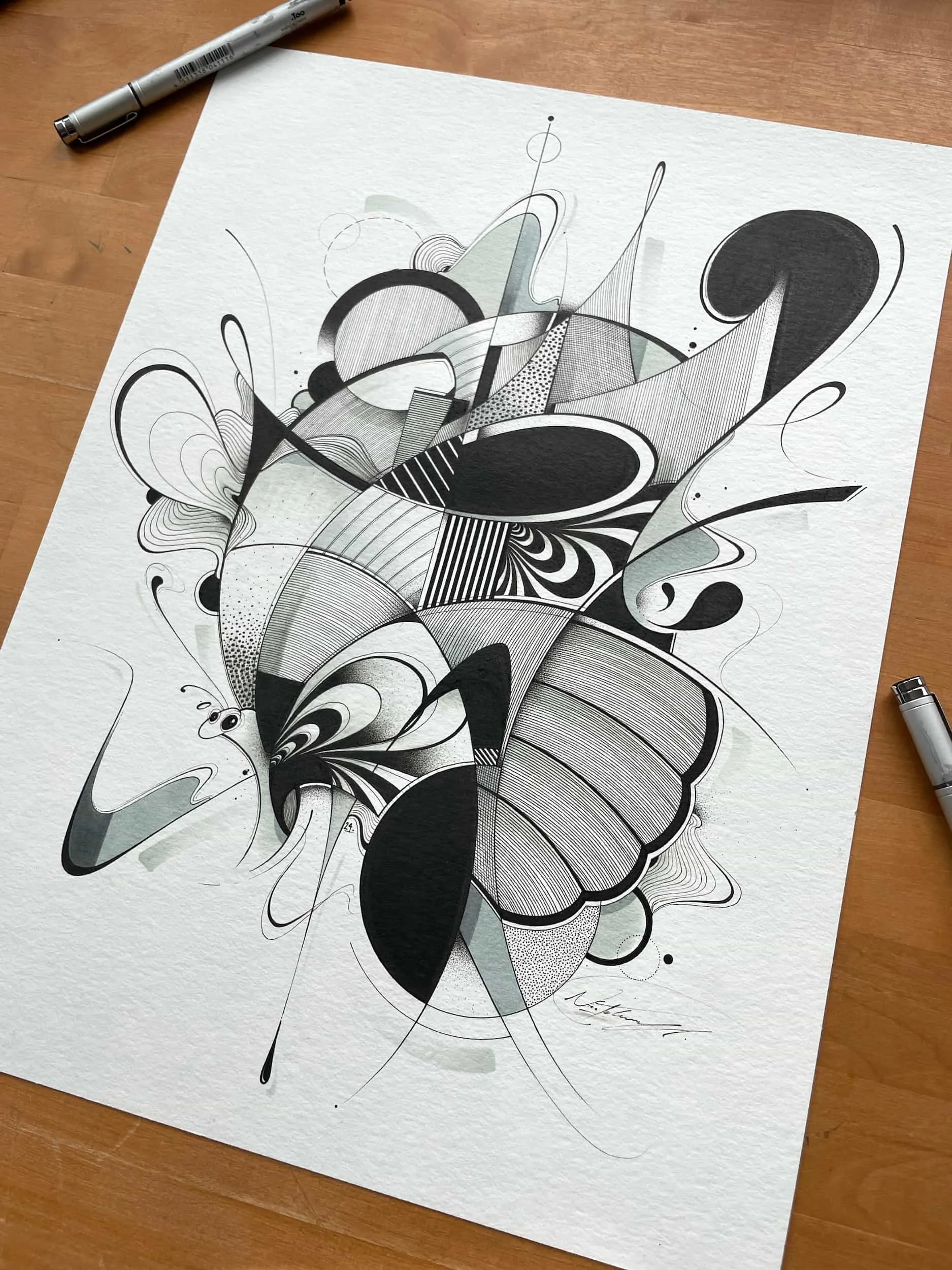 A black and white abstract geometric drawing on textured paper, with various curving and straight lines, circles, and shapes, partially colored in light green and gray, on a wooden surface with two silver pens nearby.