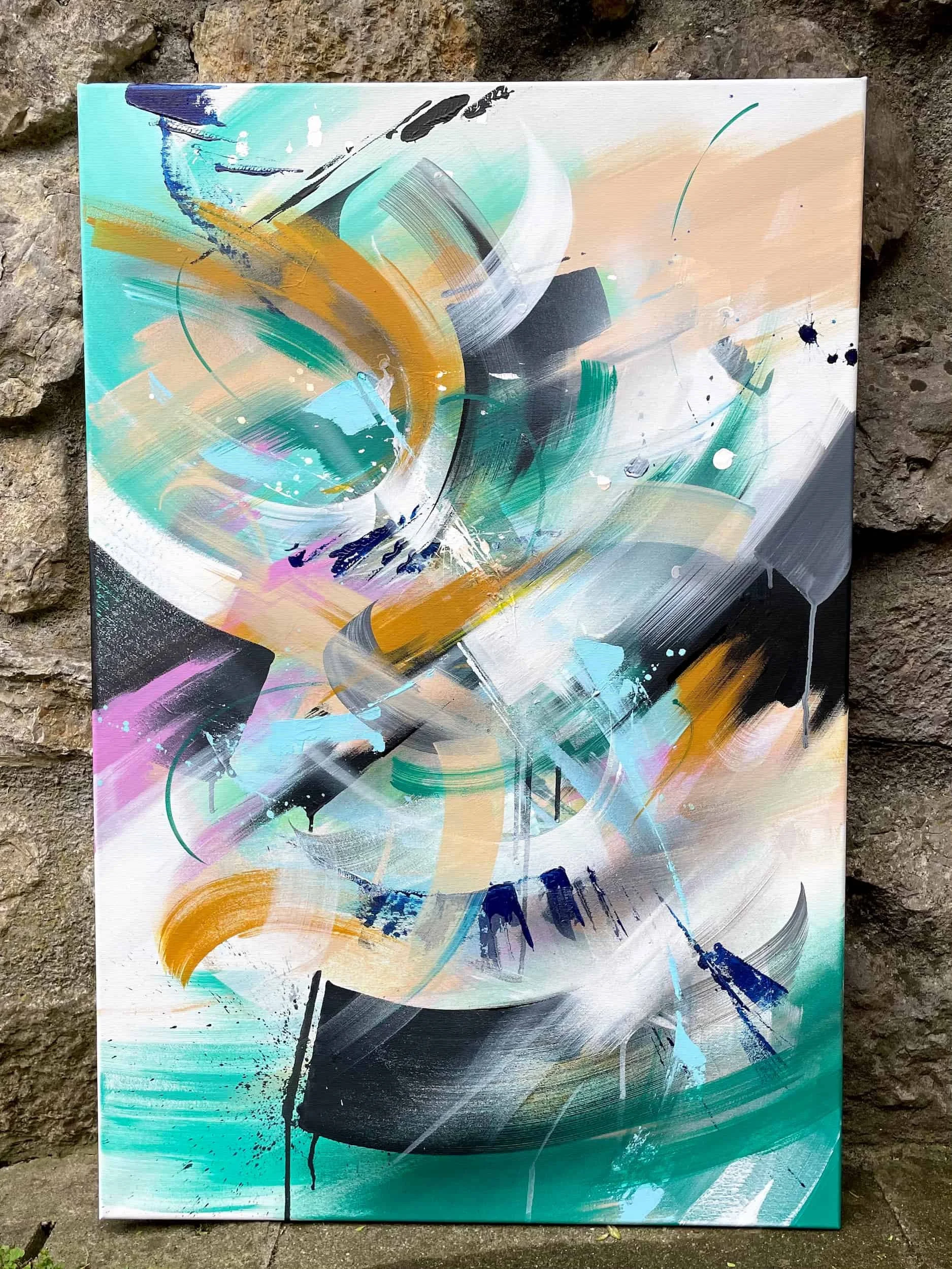Abstract painting with swirling brushstrokes of blue, black, white, peach, and gold on canvas, set against a stone wall.