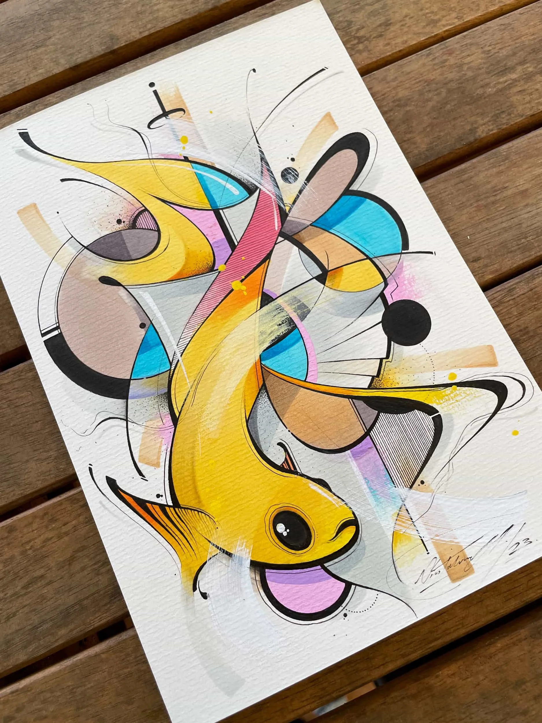 Vibrant abstract graffiti art featuring a yellow fish with a playful expression, surrounded by colorful geometric shapes, splashes, and black fine lines on textured white paper placed on a wooden surface.