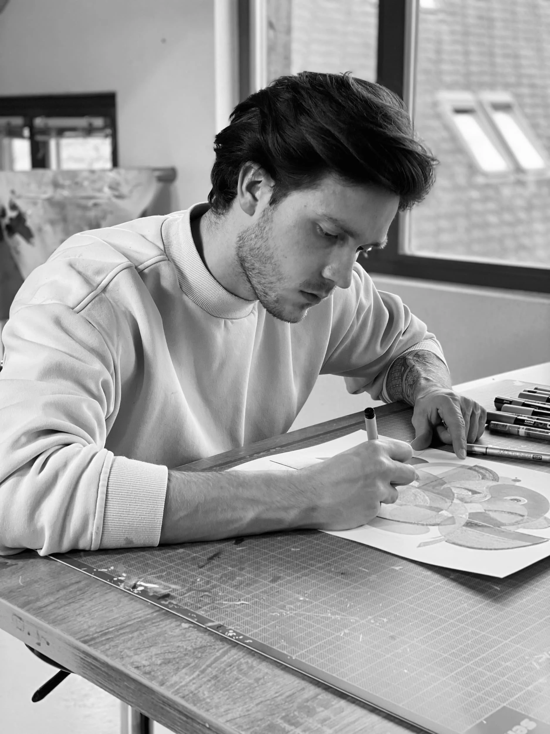 Man drawing or coloring on a piece of paper with markers on a table, with a window in the background.