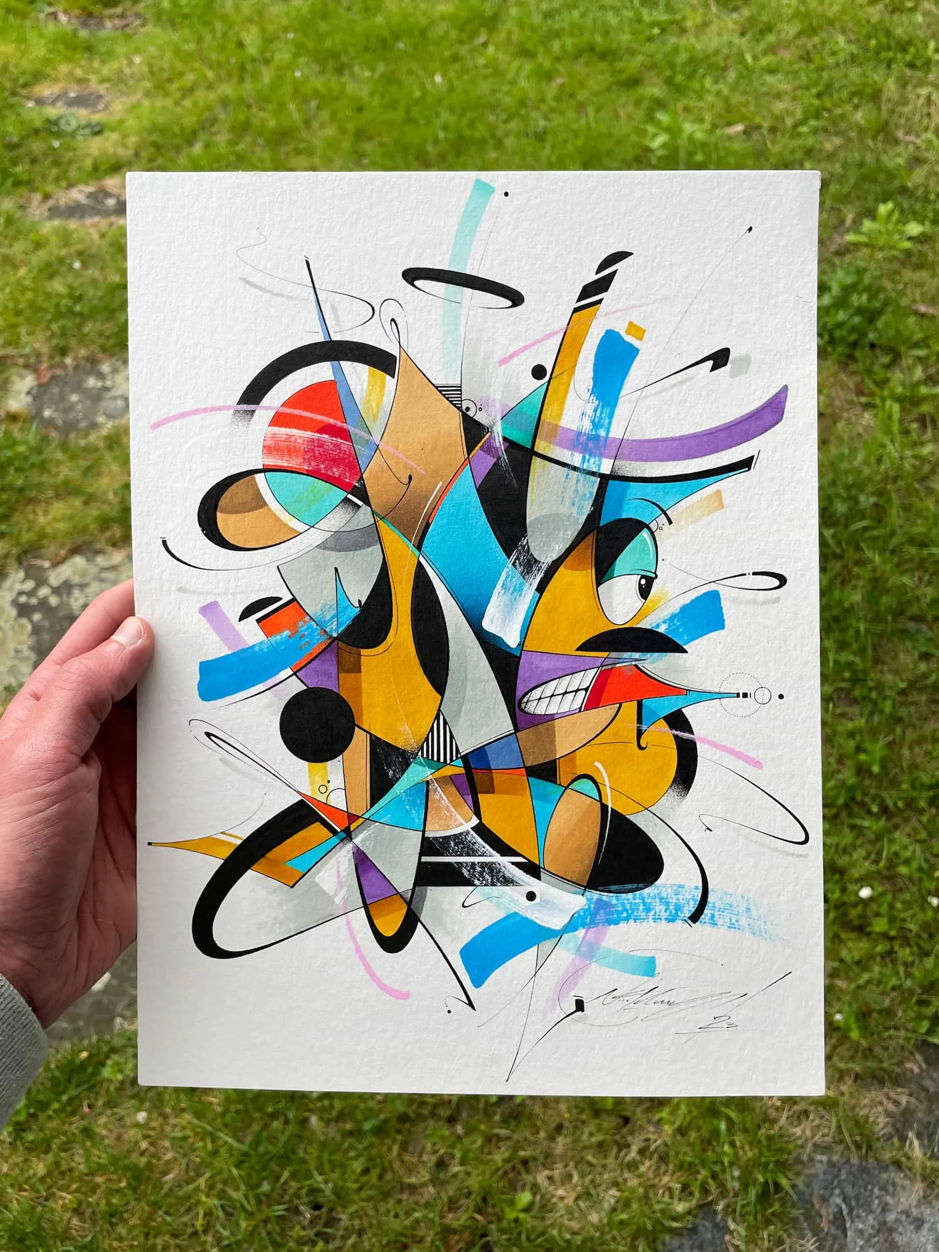 Abstract colorful painting with black, blue, yellow, purple, and white shapes and lines, held in a person's left hand outdoors.